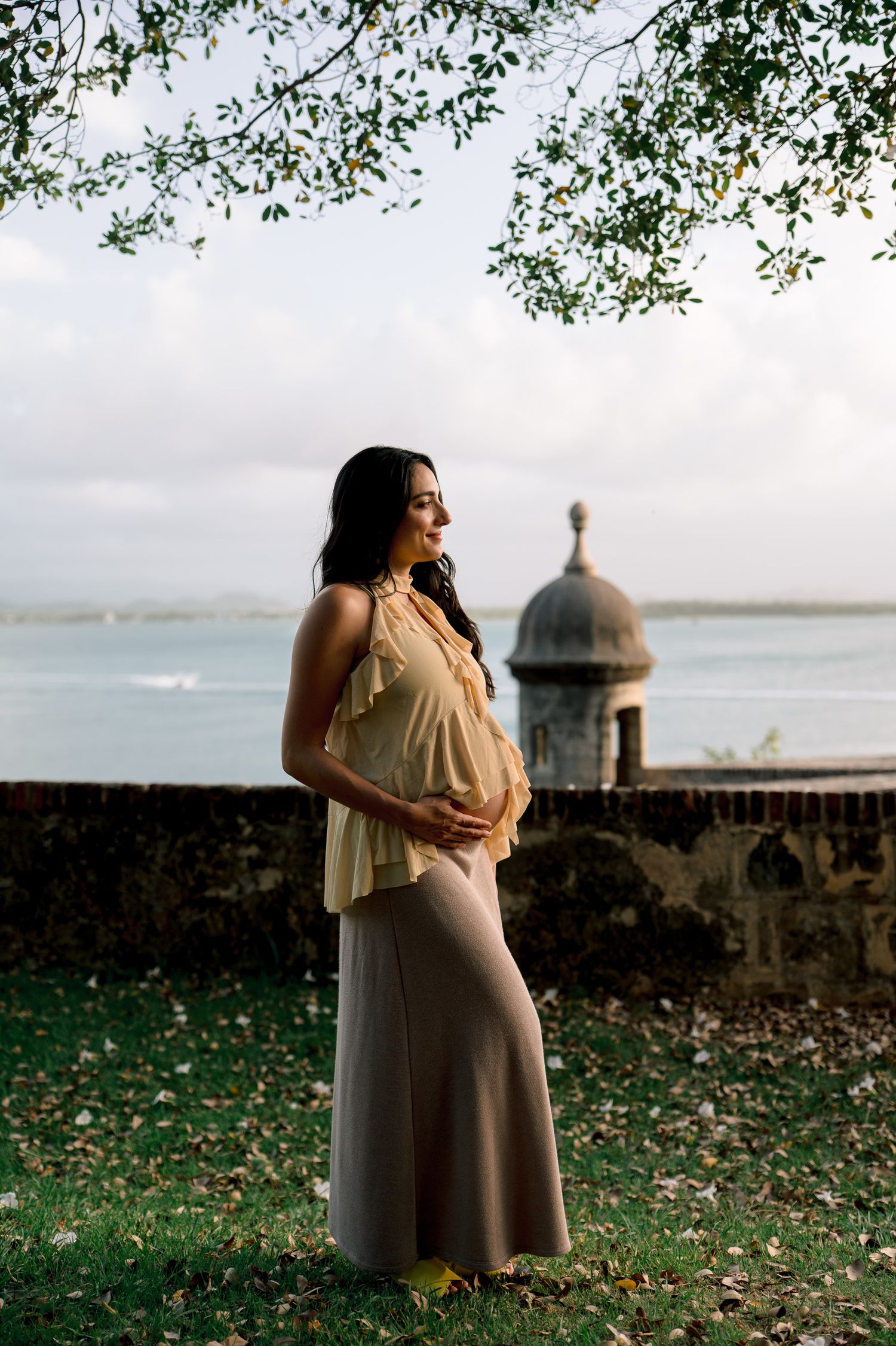 Pregnant woman in yellow top and tan skirt cradling her belly near a historic stone wall by the ocean.