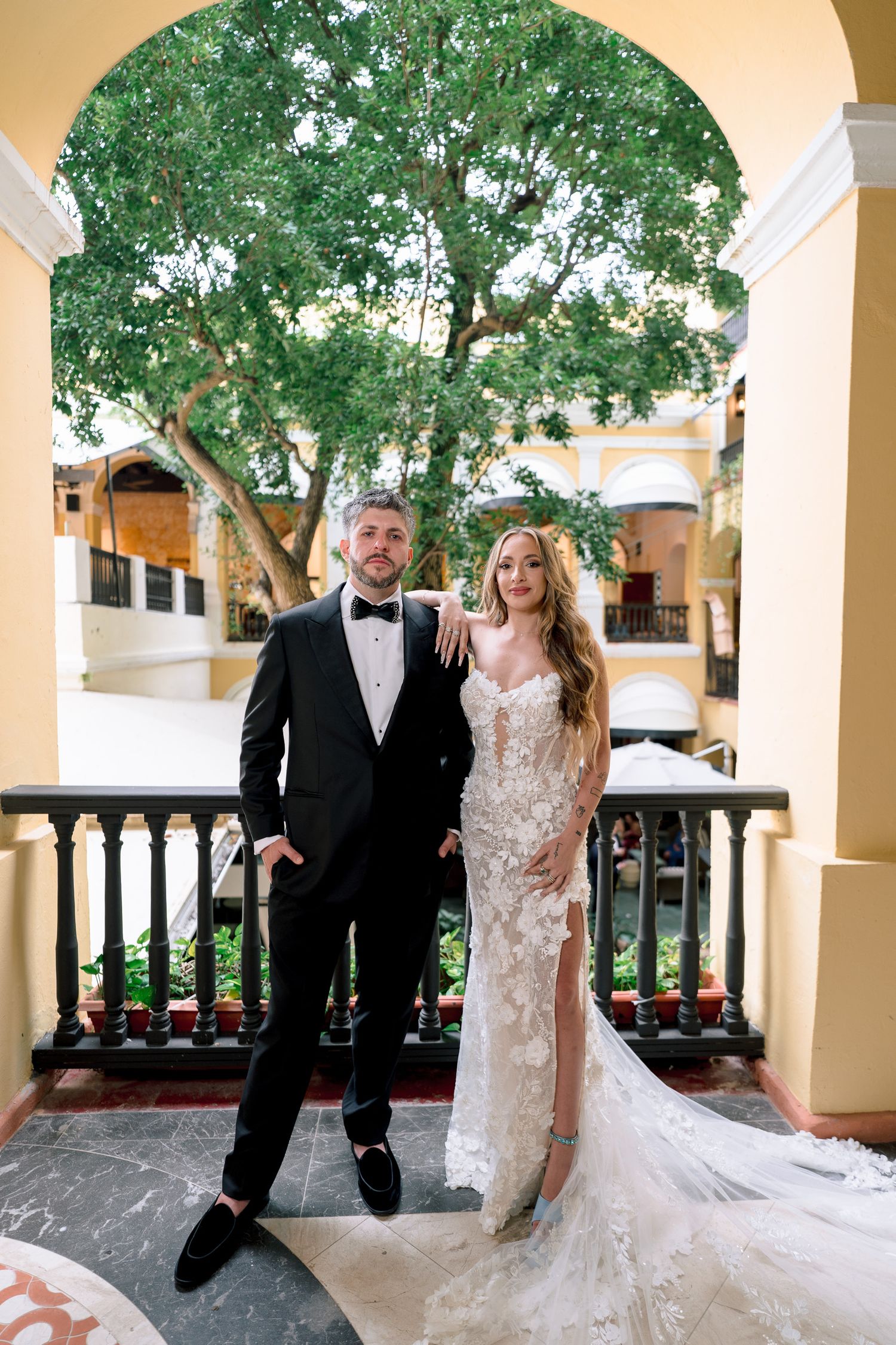 Bride in lace gown and groom in black tuxedo pose on balcony with arched columns and lush green tree backdrop.