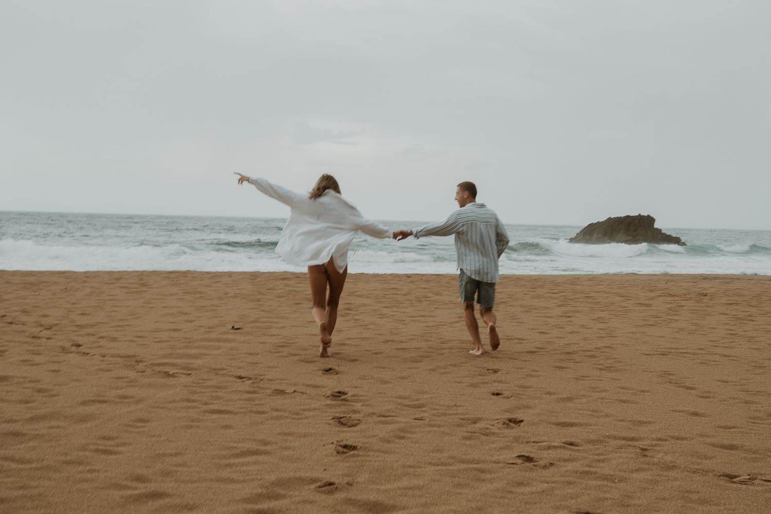 Joyful couple running hand in hand along a sandy beach with ocean waves behind them.