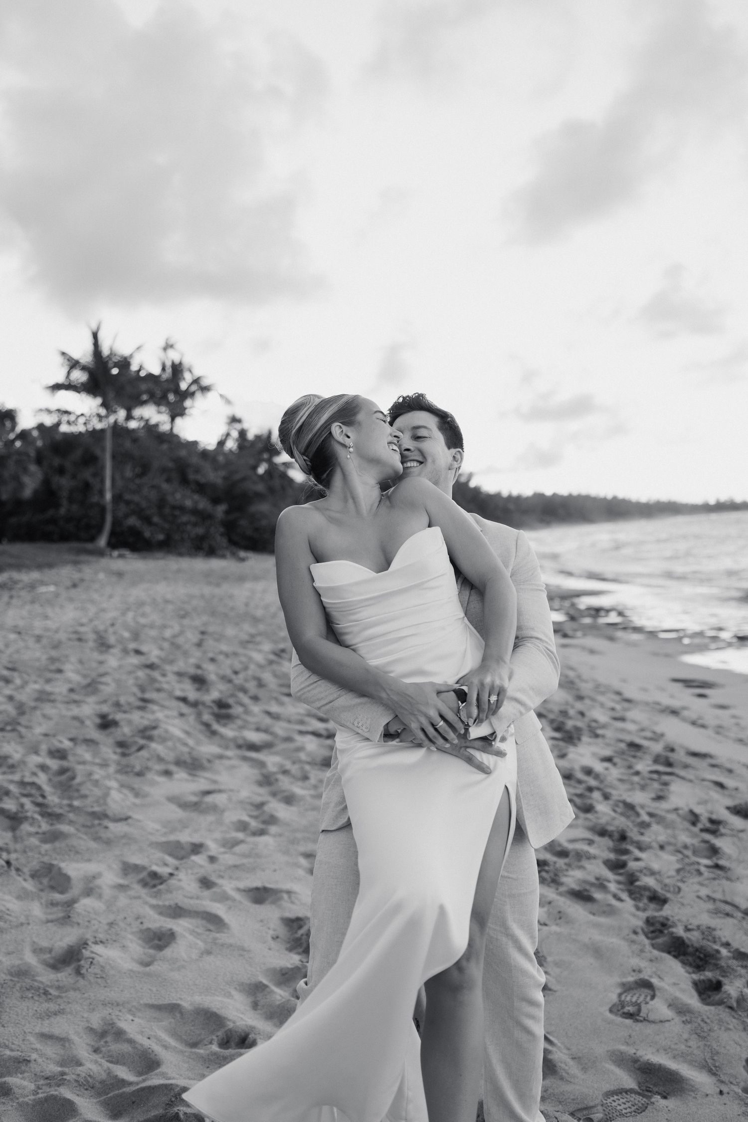 Black and white photo of bride and groom embracing on a tropical beach at sunset.