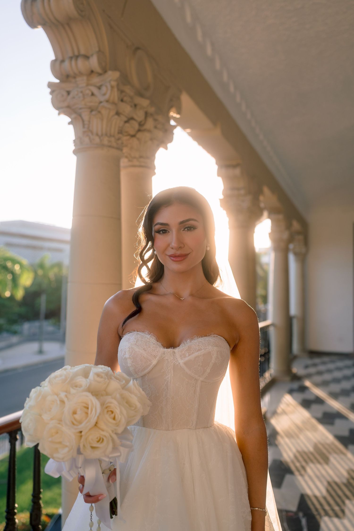 Bride in strapless lace wedding gown holds white rose bouquet on sunlit columned balcony.