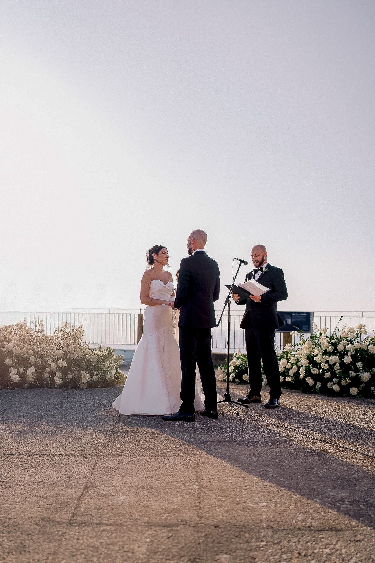 Bride and groom exchange vows outdoors with officiant at elegant seaside wedding ceremony.