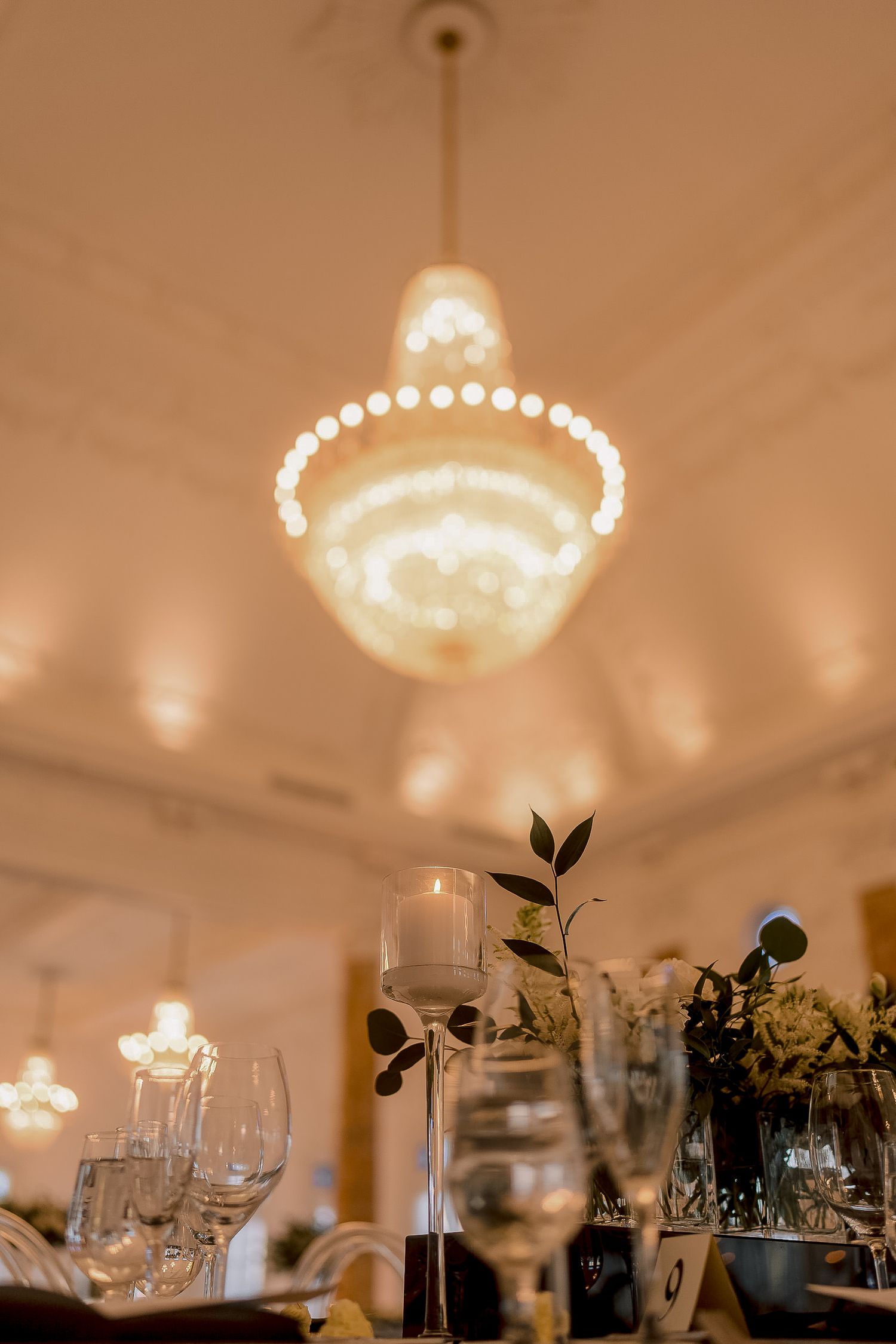 Elegant wedding reception table setting with glassware and floral centerpieces beneath a glowing chandelier.