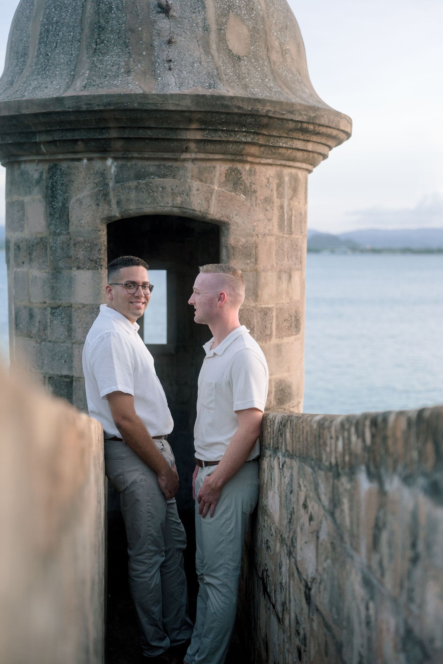 Two men in white shirts share a romantic moment at a historic stone fort turret overlooking the ocean.