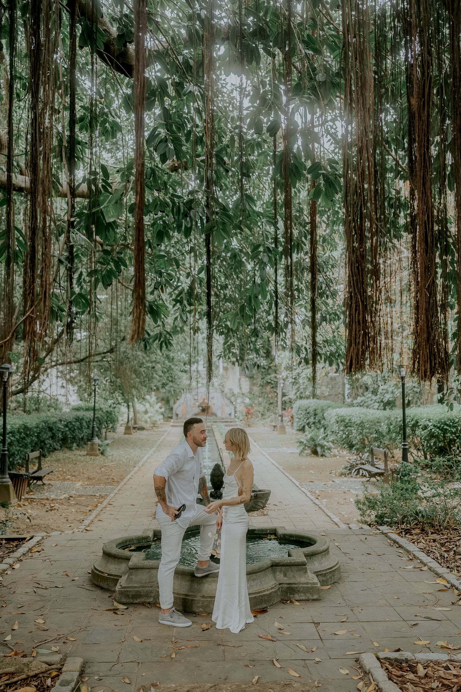 Couple in white attire standing by a stone fountain surrounded by tall trees in a lush garden.