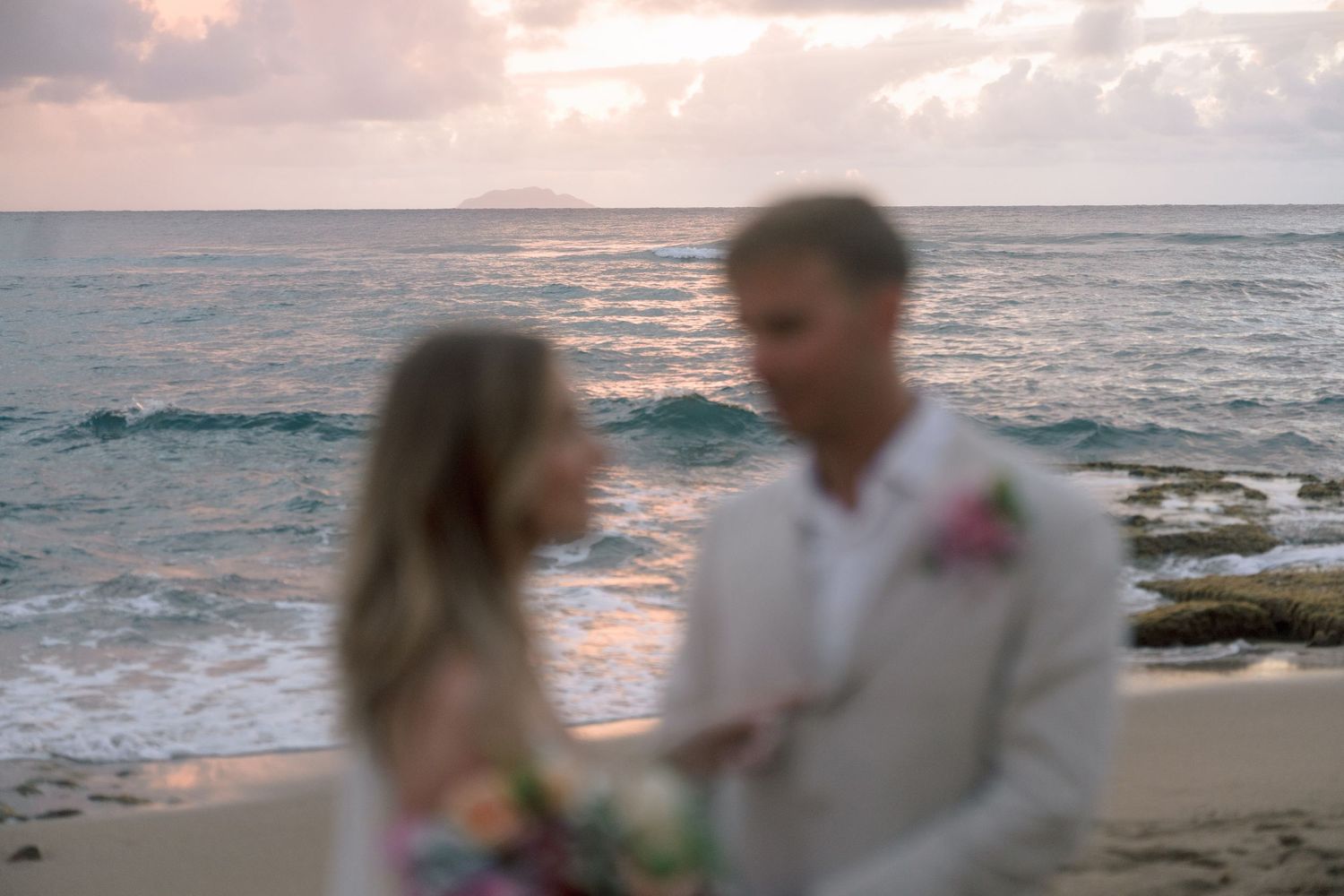 Blurred romantic photo of a bride and groom facing each other on a beach at sunset.