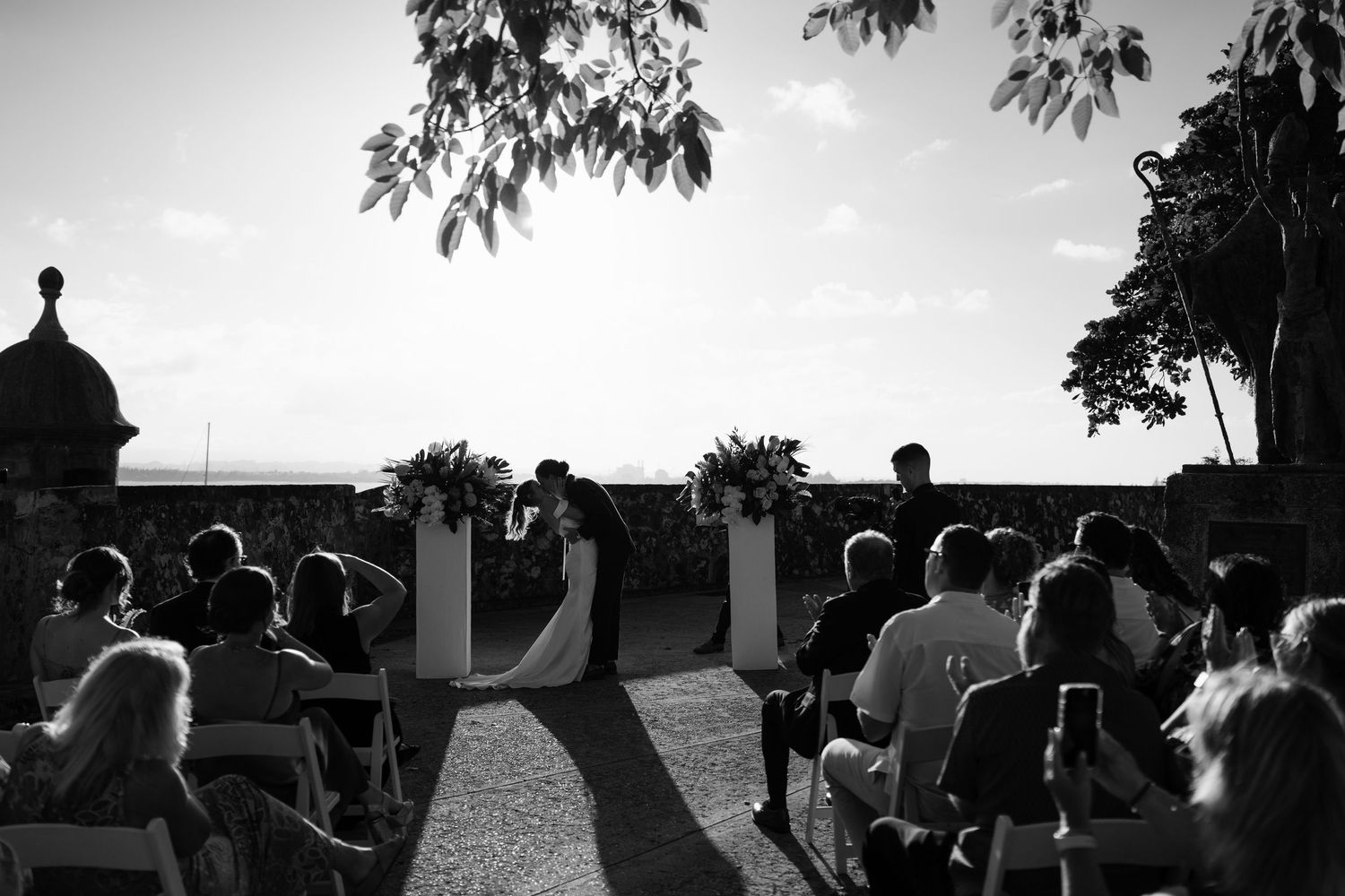 Black and white outdoor wedding ceremony with guests seated, couple at altar, ocean view backdrop.
