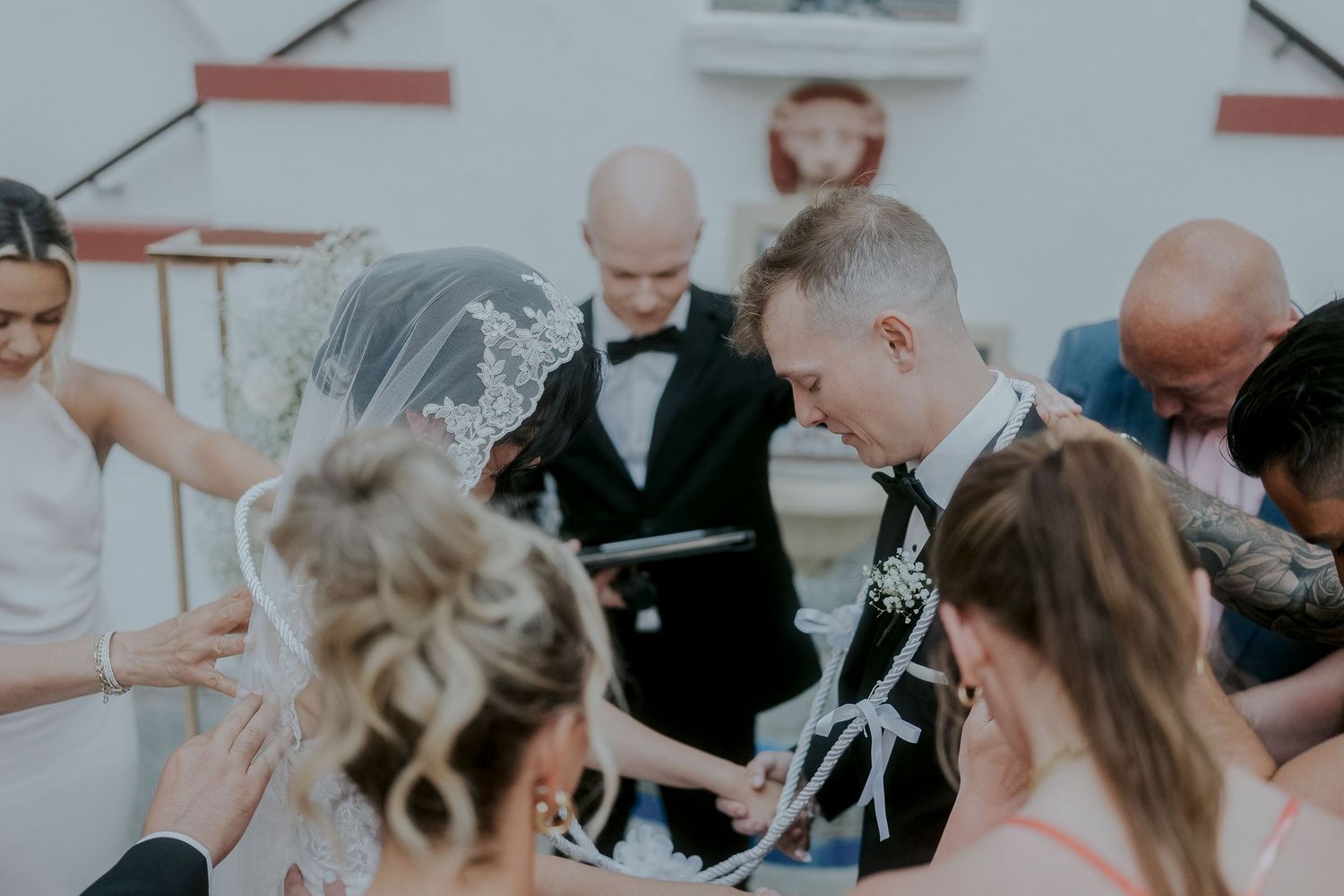 Wedding couple in formal attire surrounded by guests in a circle, sharing a prayer during an outdoor ceremony.
