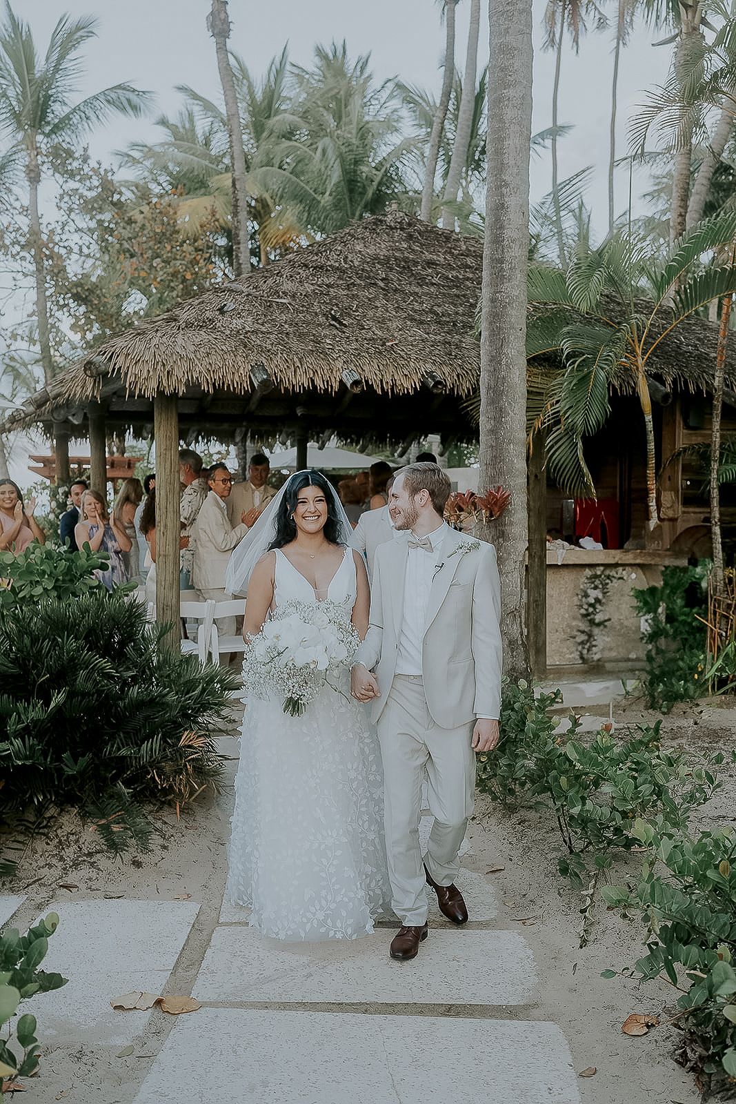 Bride and groom walk hand in hand down sandy aisle at tropical beach wedding with palm trees and thatched gazebo.