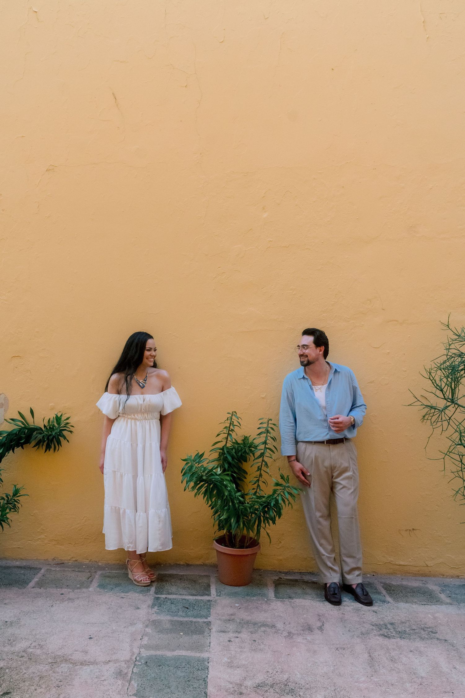 A couple stands against a vibrant yellow wall with potted plants, woman in white dress, man in blue shirt holding a drink.