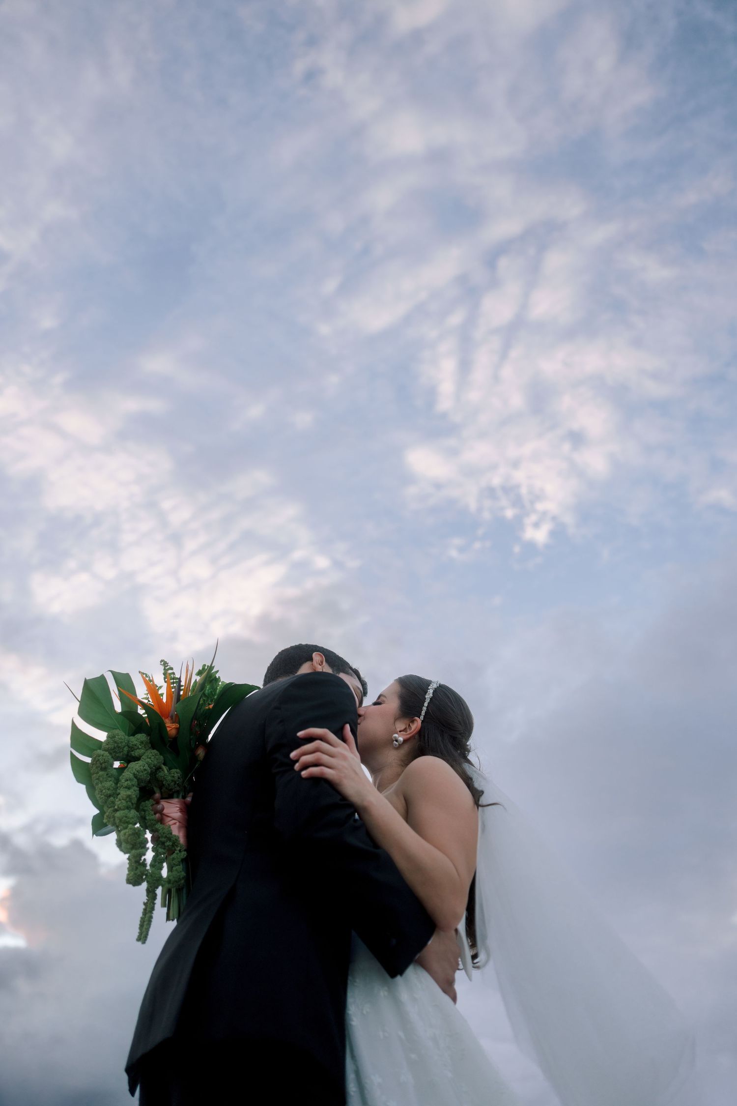 Newlyweds share a kiss beneath a dramatic cloudy sky, bride holding a lush tropical green and orange bouquet.