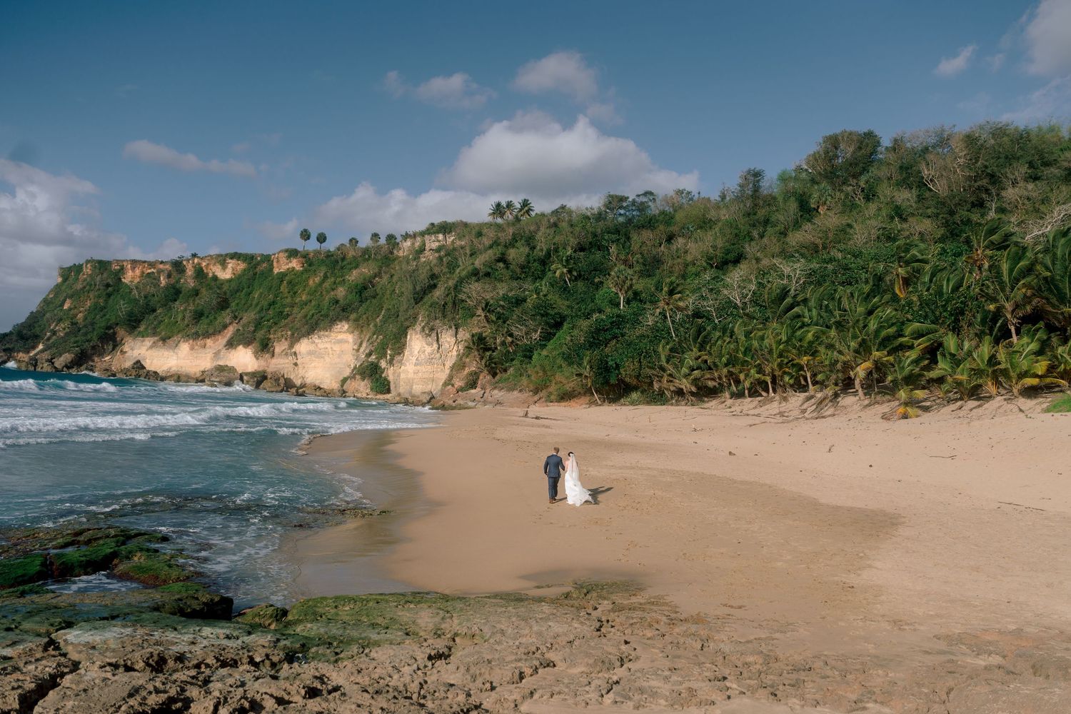 Bride and groom walking on a secluded tropical beach with lush green cliffs and golden sand.