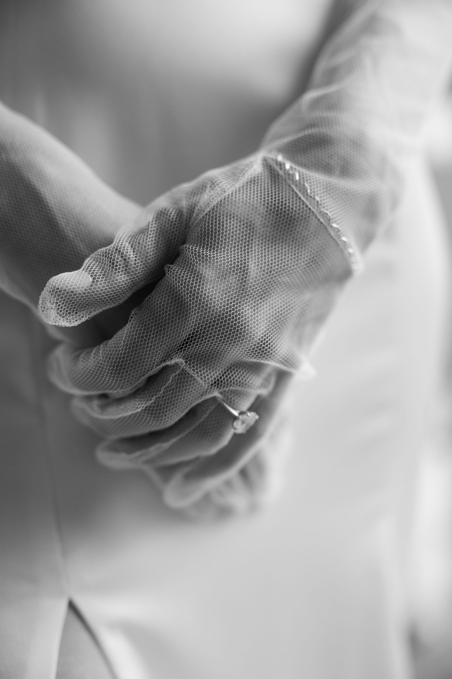 Black and white close-up of bride's hands wearing lace gloves and diamond engagement ring on wedding day.