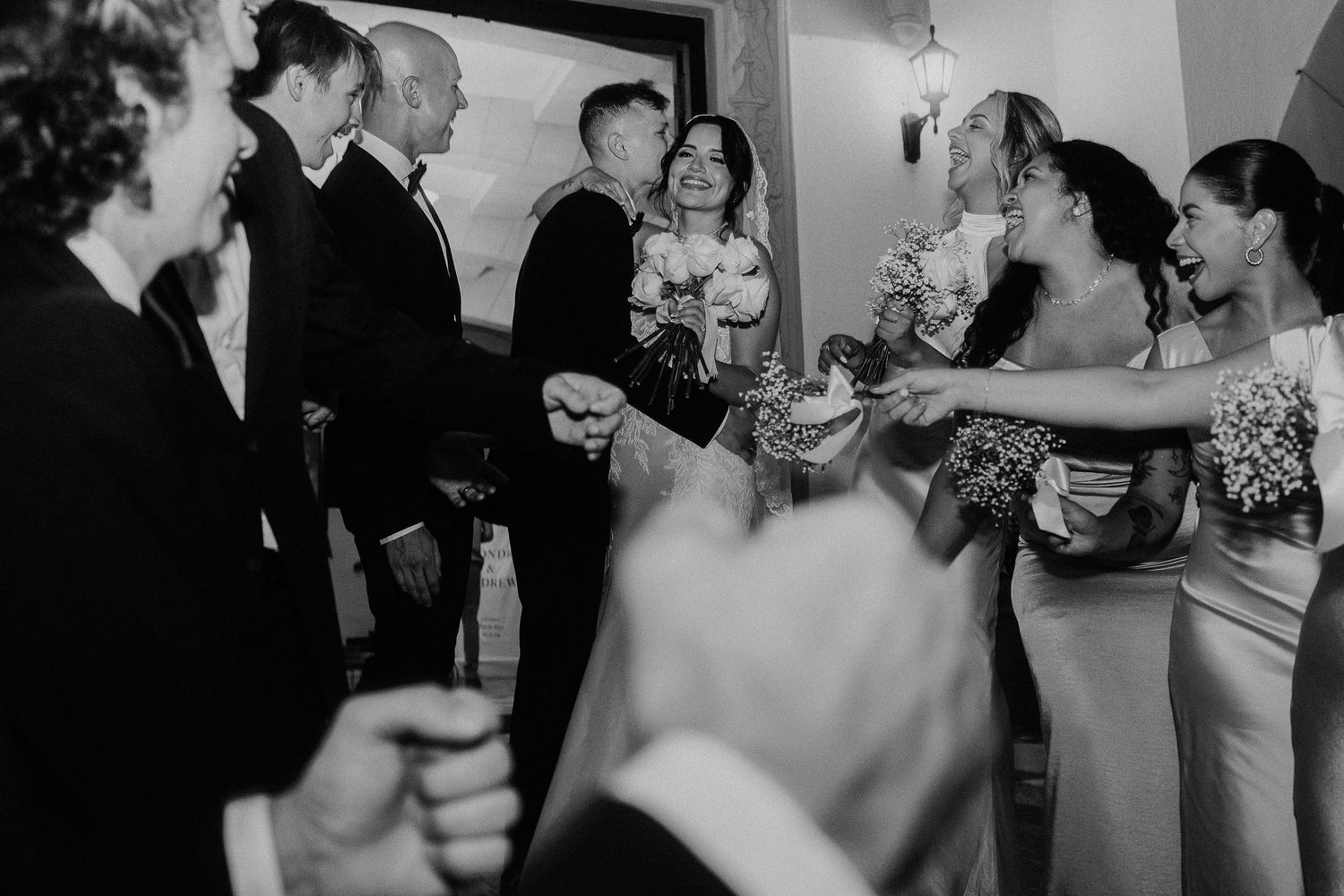 Black and white wedding photo of bride, groom, and bridesmaids celebrating with guests in elegant venue.