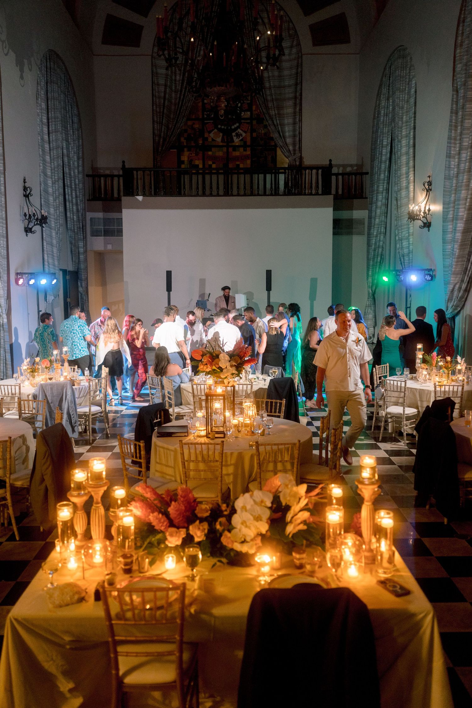 Candlelit wedding reception with guests dancing in grand venue featuring checkered floors and arched windows.