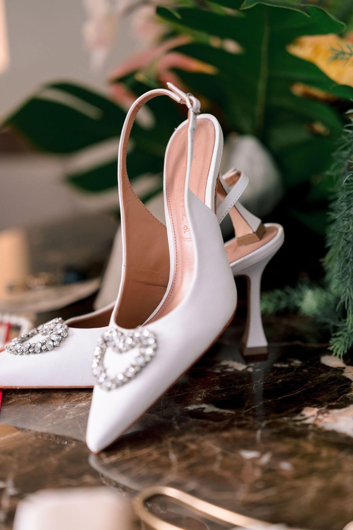 White satin slingback wedding heels with crystal heart buckle detail on marble surface with greenery.