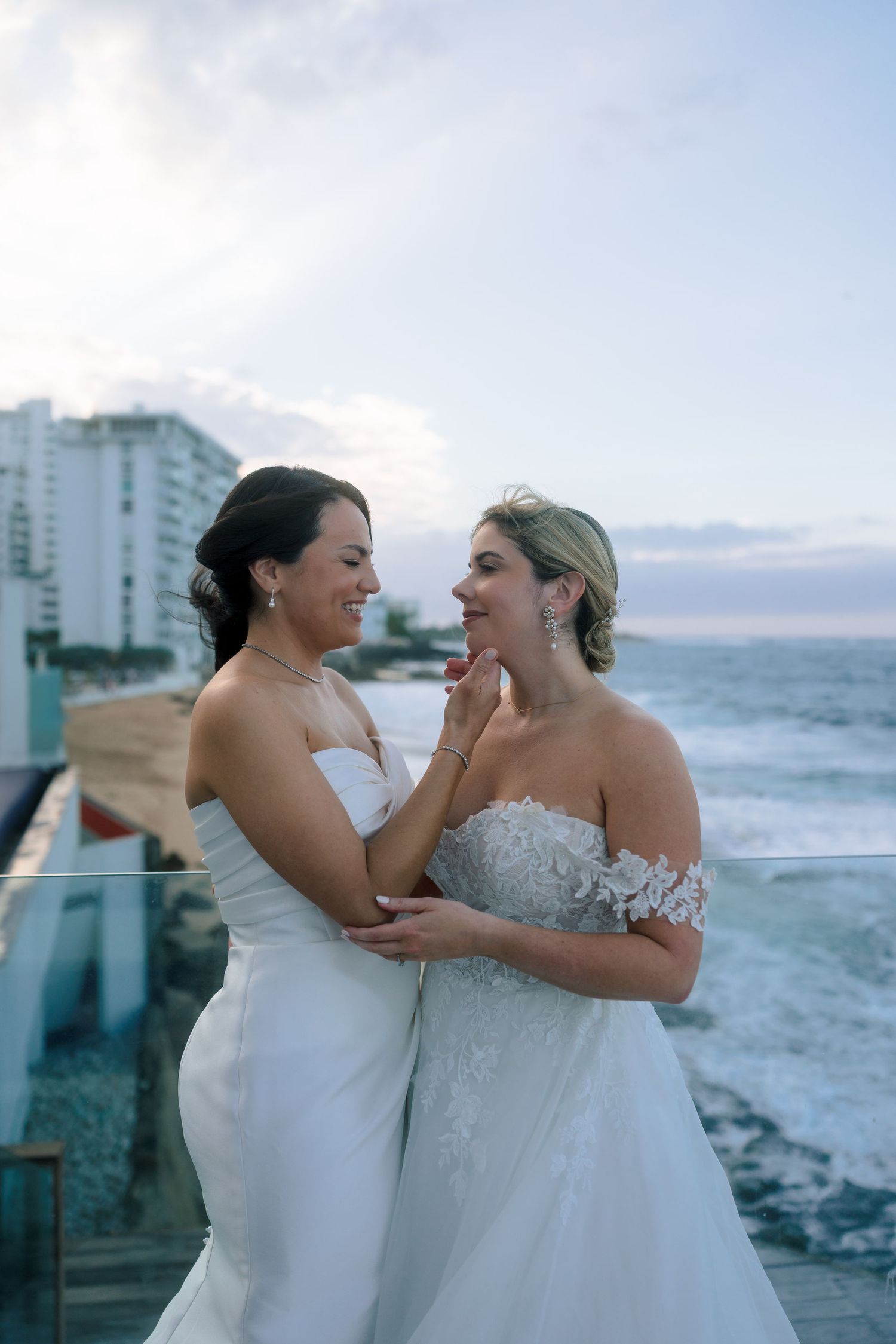 Two brides in white wedding gowns embrace on a beachfront balcony, smiling at each other with ocean waves behind them.