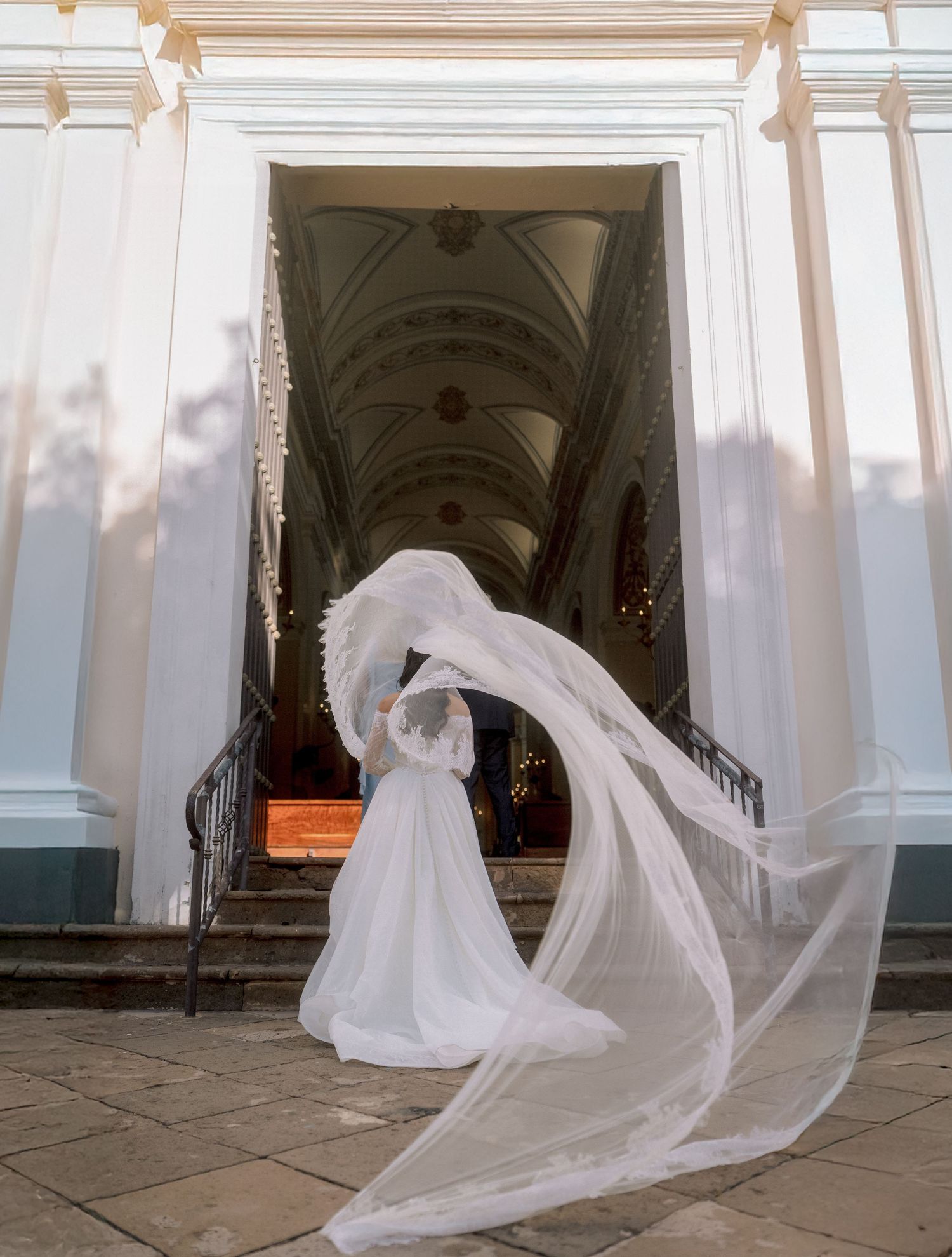 Bride in flowing white gown and veil billowing dramatically at ornate church entrance with arched ceiling.
