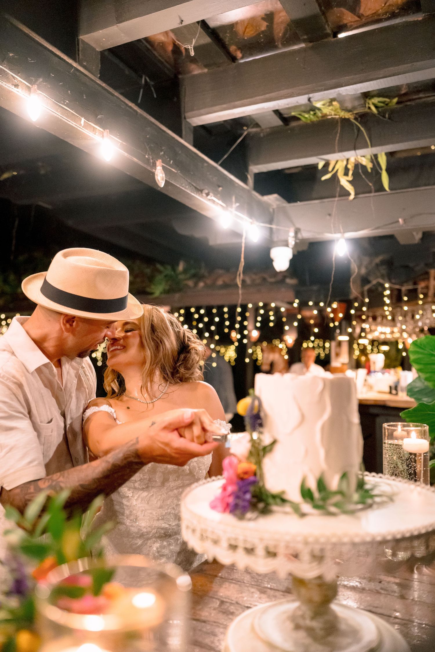 Couple sharing a kiss while cutting their wedding cake under string lights at a romantic outdoor reception venue.