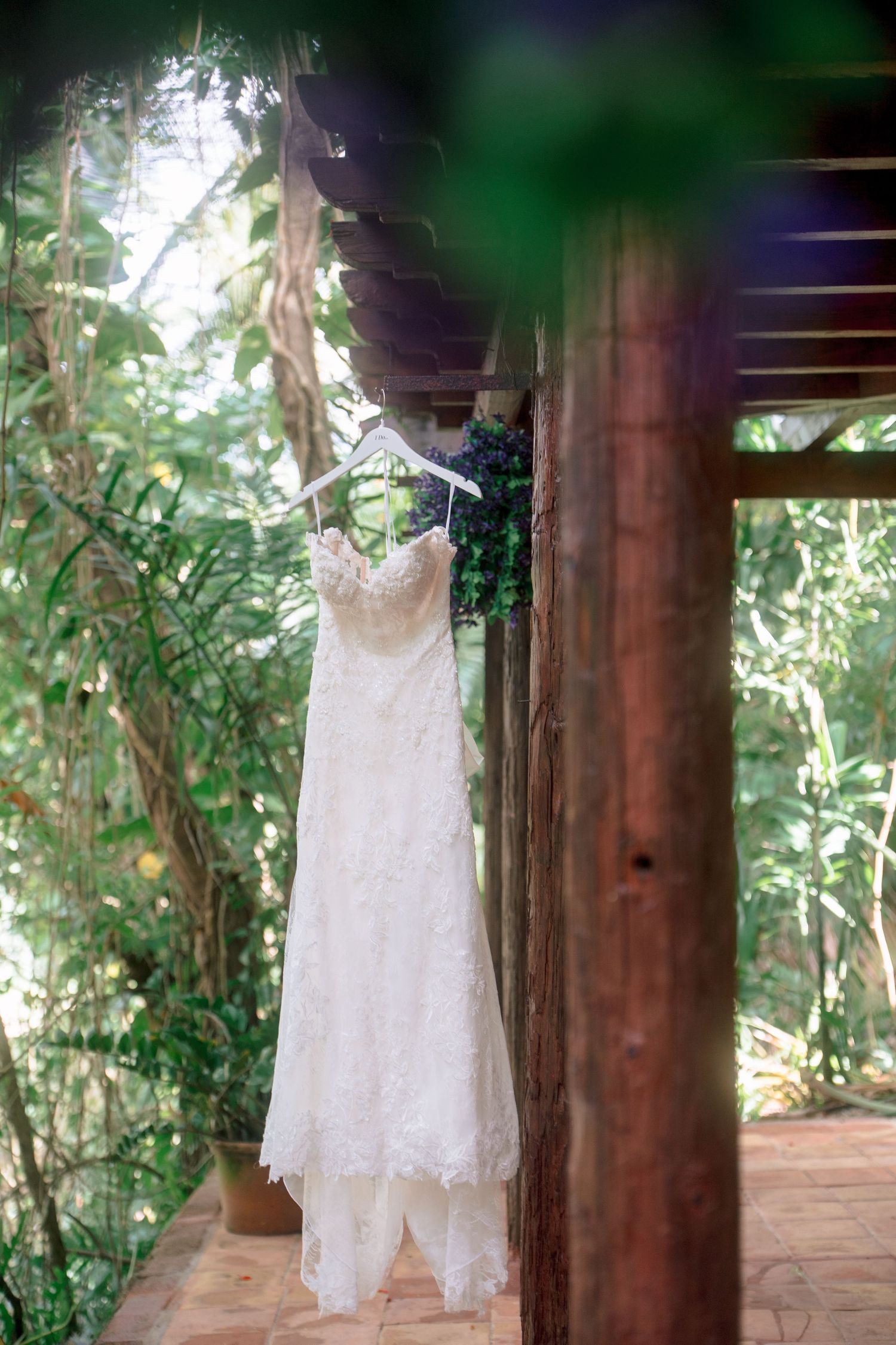 A white lace wedding dress hanging on a wooden beam surrounded by lush green tropical foliage outdoors.