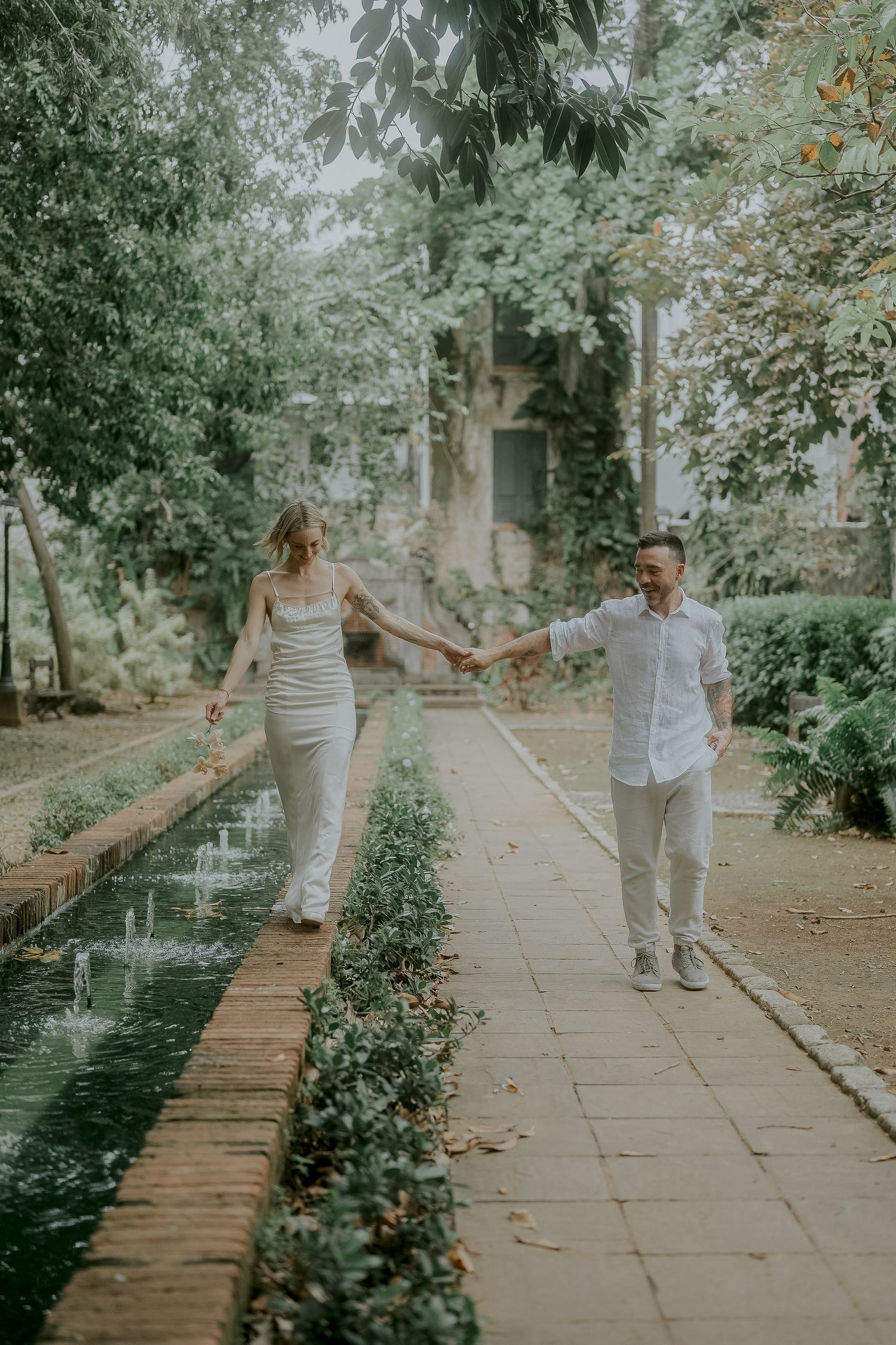 Couple holding hands walking along a garden path beside a fountain, dressed in white for an outdoor engagement photo.