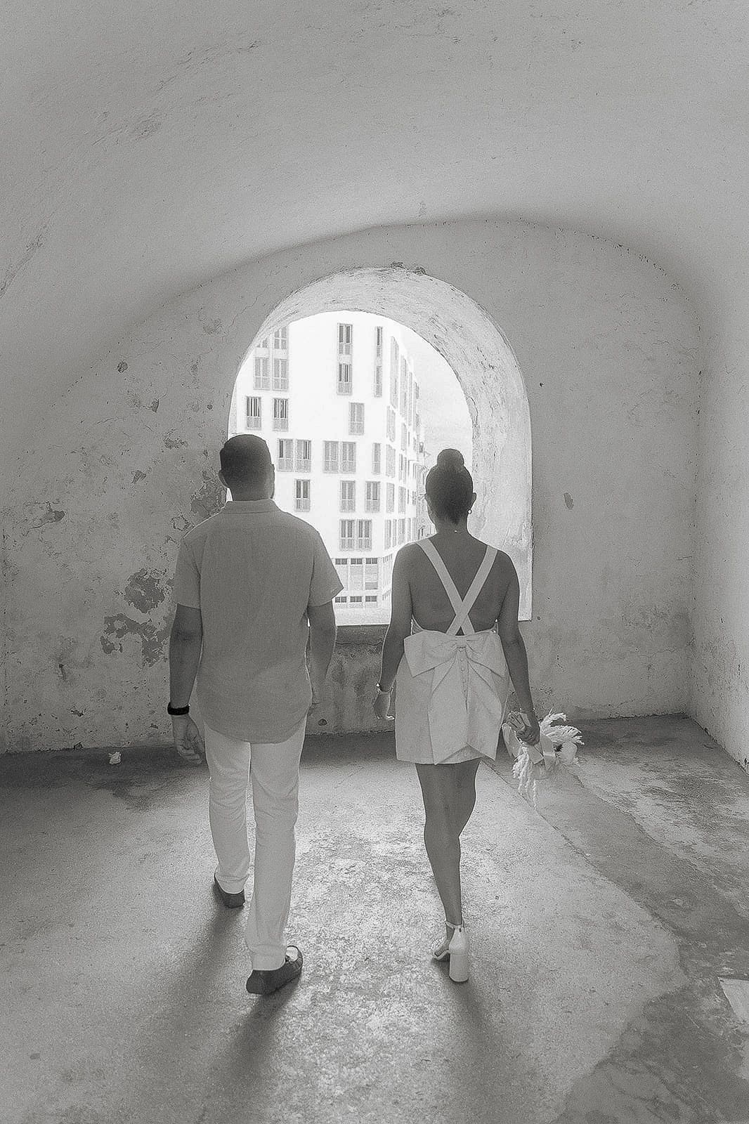 Black and white photo of a couple walking hand in hand through an arched tunnel toward a bright opening.