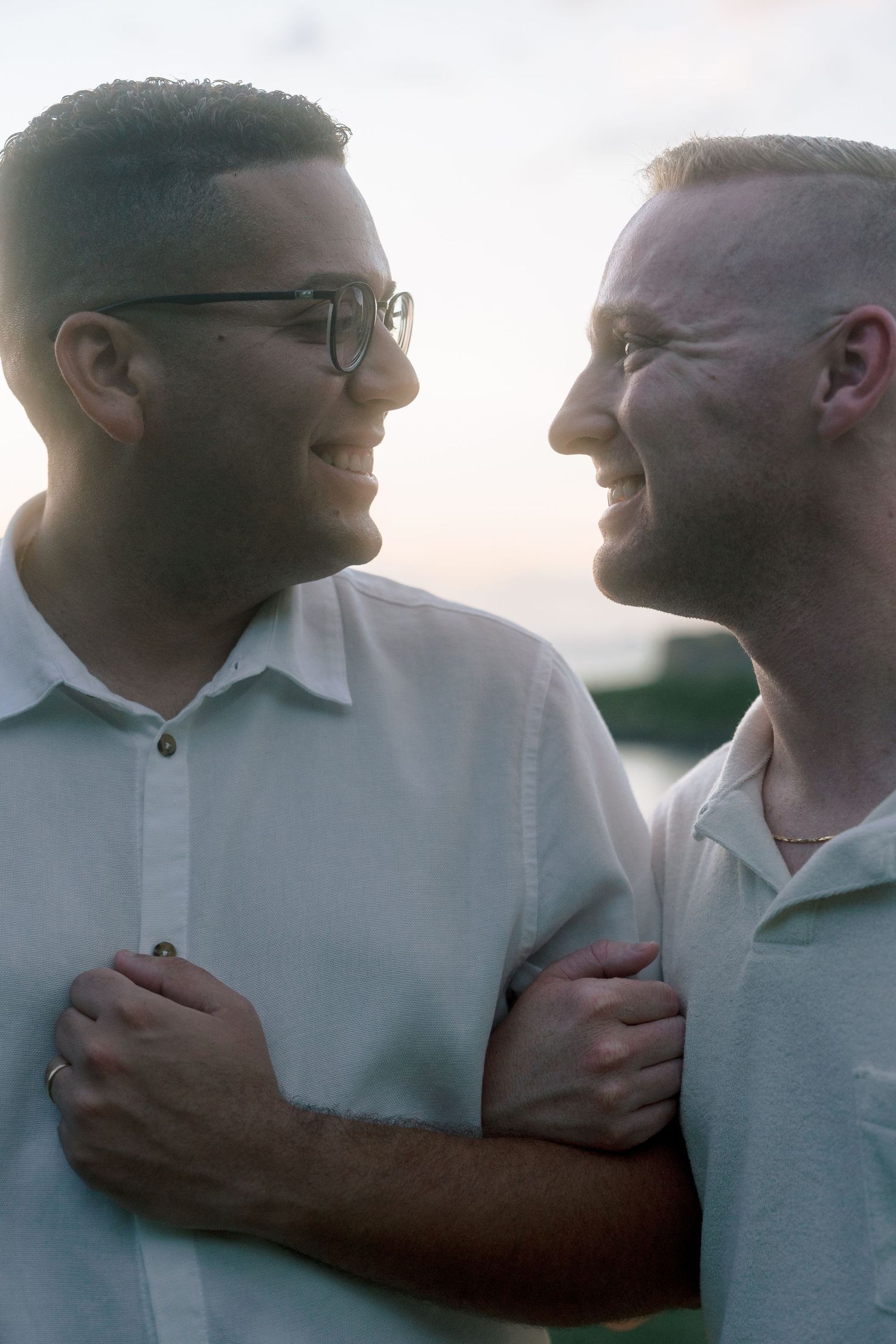 Two smiling men embrace closely, sharing a joyful moment with soft sunset light behind them.