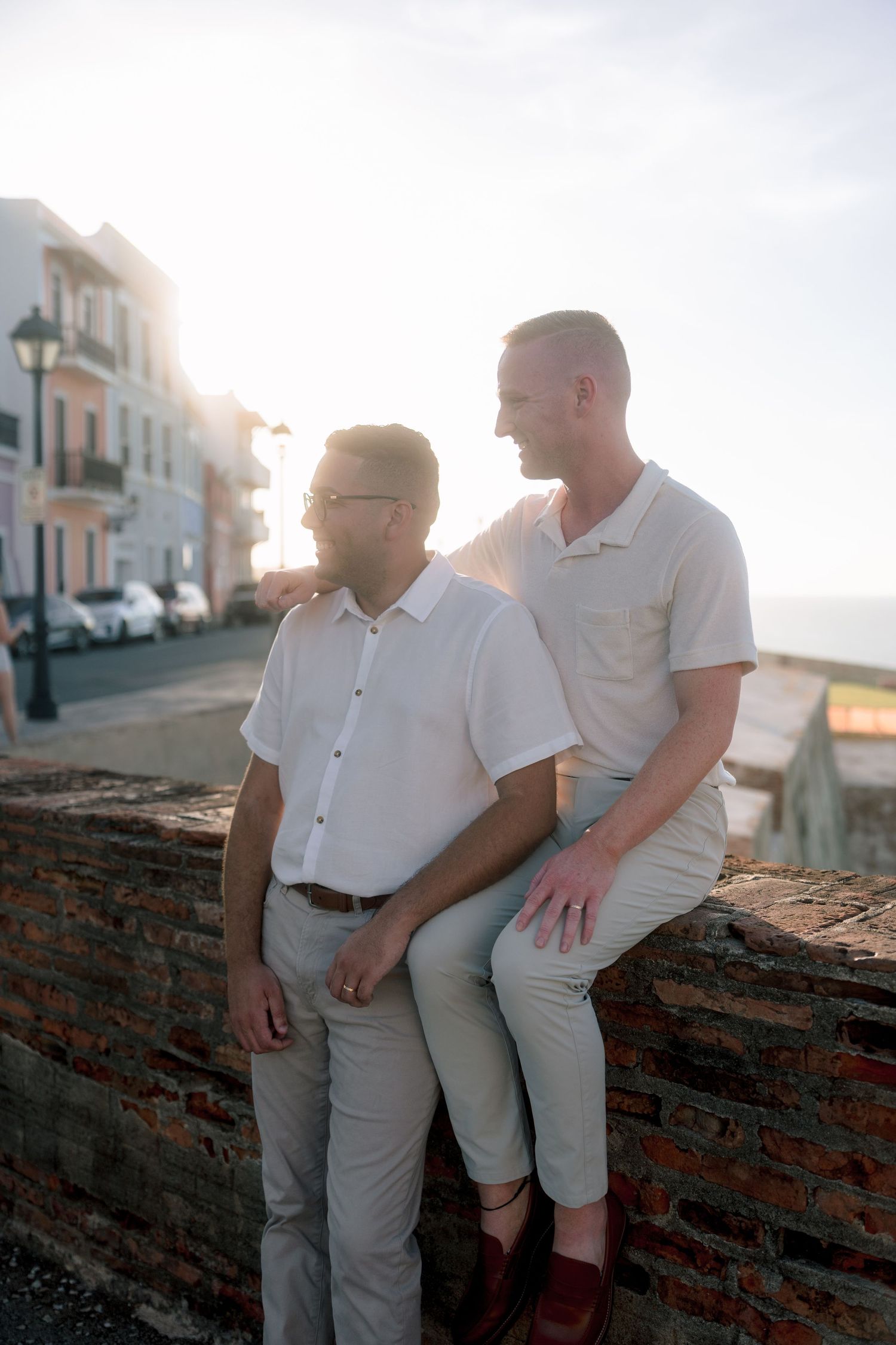 Two men in white shirts sit on a brick wall at sunset in a colorful coastal town street.