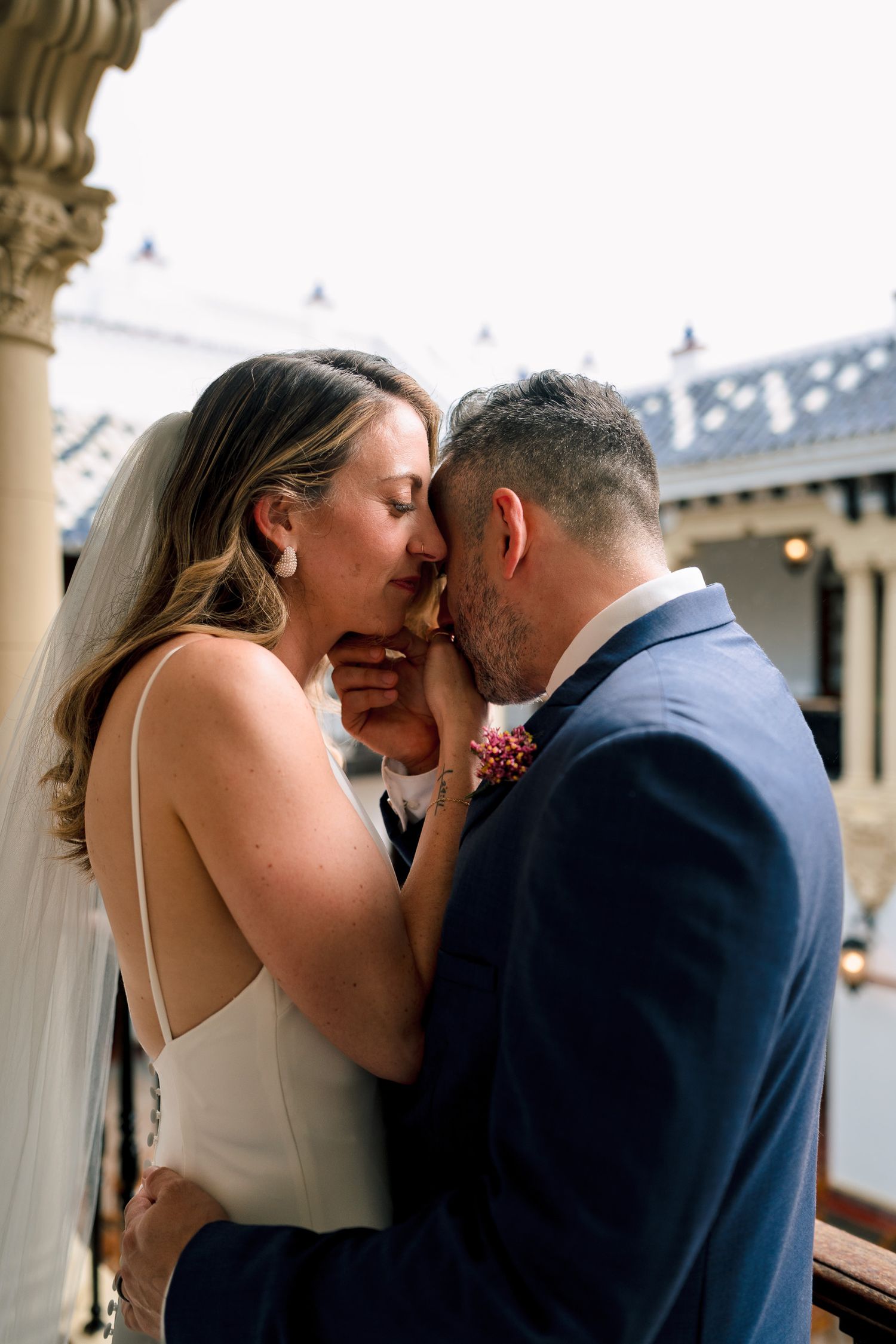 A bride and groom share an intimate moment on a balcony, the groom kissing her hand tenderly.