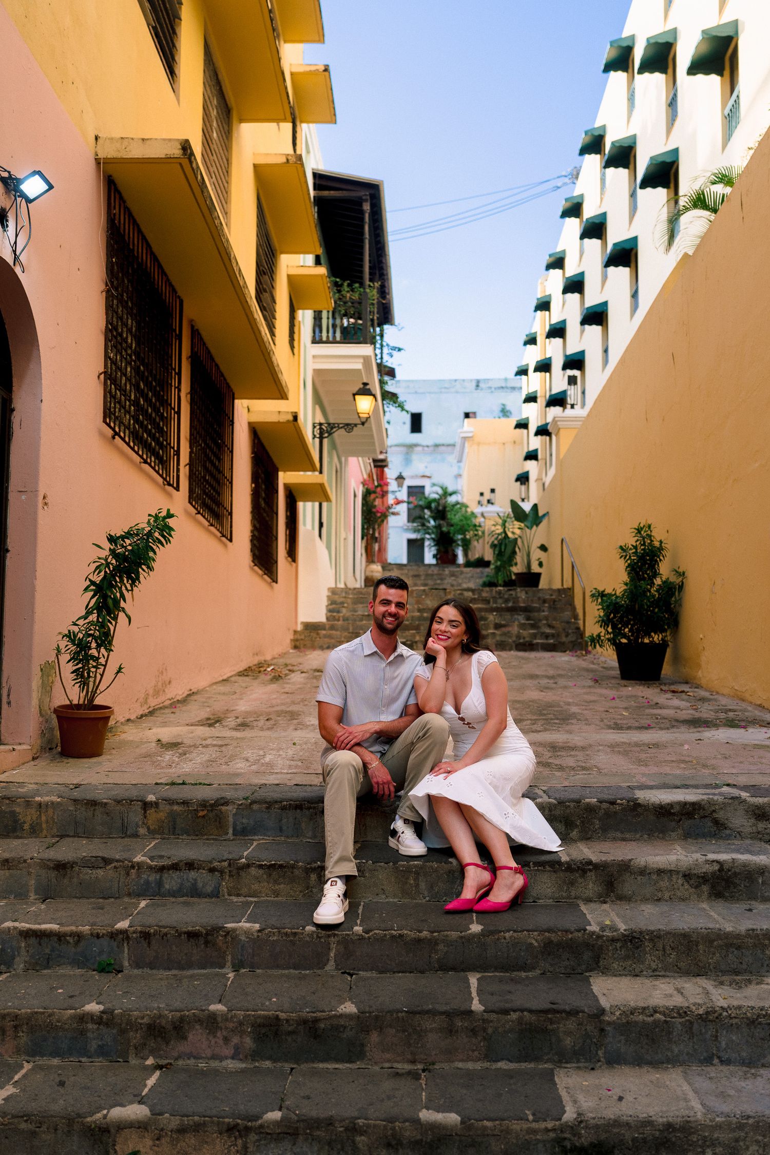 Couple sitting on colorful cobblestone steps in Old San Juan, Puerto Rico, surrounded by vibrant yellow and pink buildings.