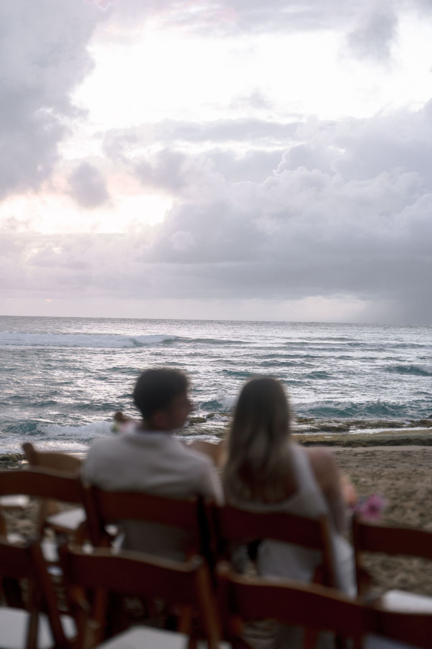 A couple sits together on wooden chairs gazing at the ocean waves under a cloudy sky at dusk.