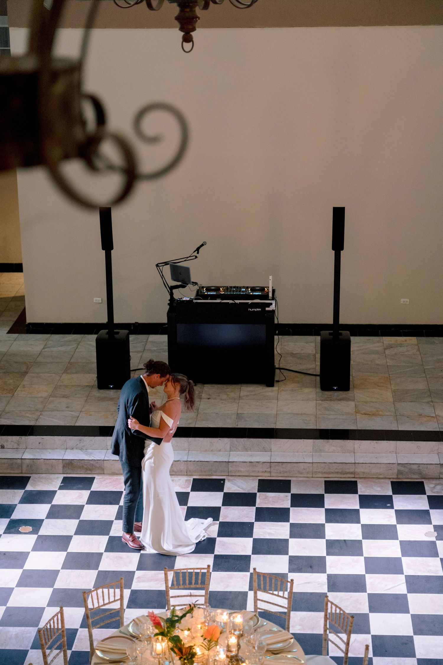 Bride and groom share first dance on black and white checkered floor with DJ setup in elegant wedding venue.