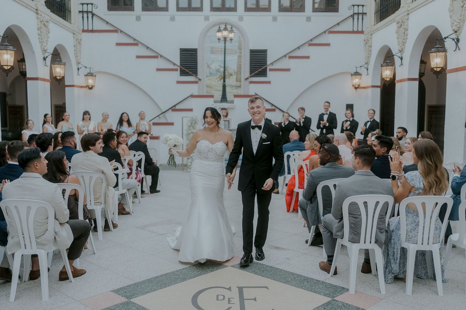 Bride and groom walk down the aisle at an elegant outdoor wedding ceremony with white chairs and arched architecture.