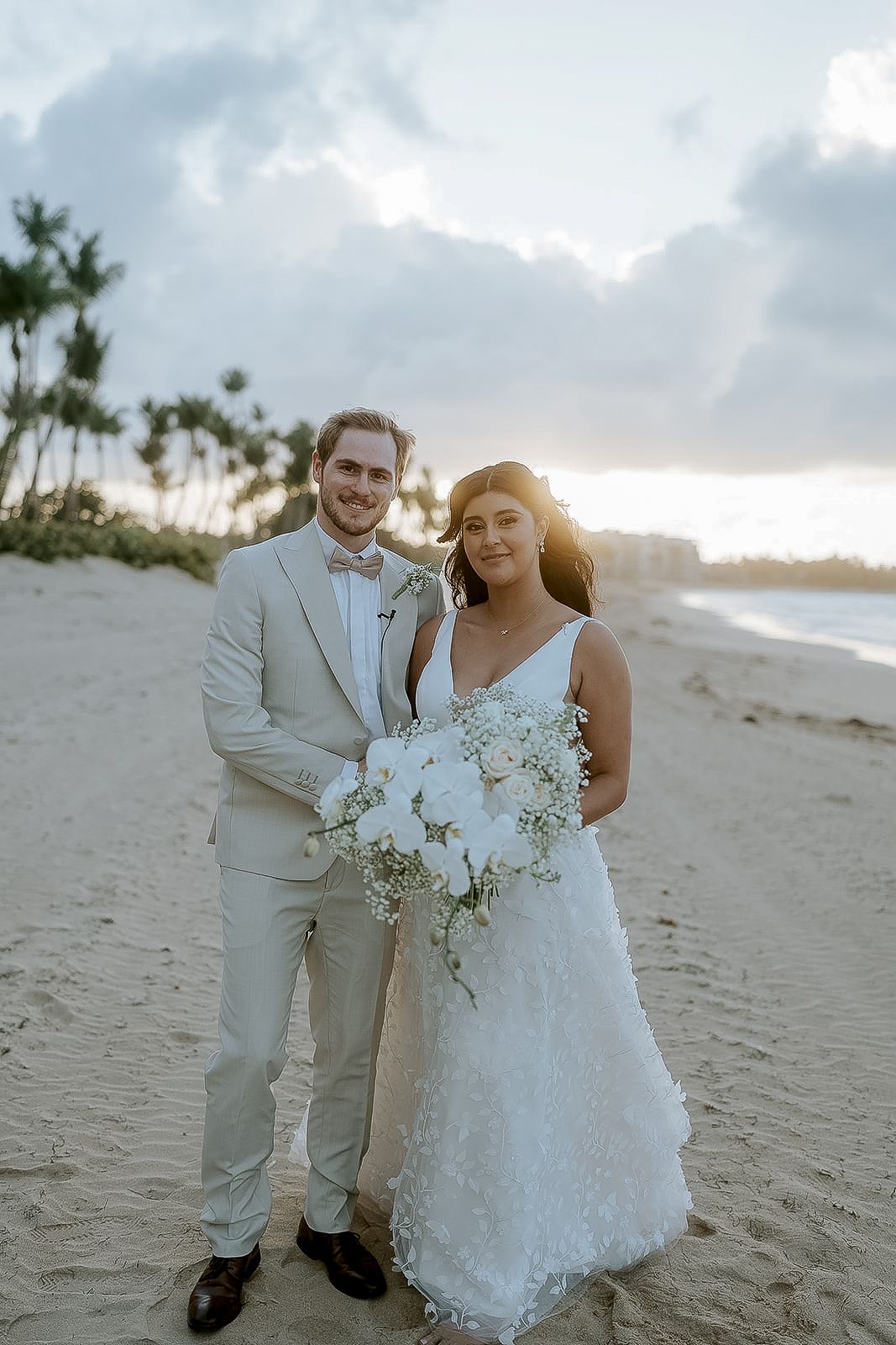 Bride and groom pose on a sandy beach at sunset, she holds a white floral bouquet, he wears a light gray suit.