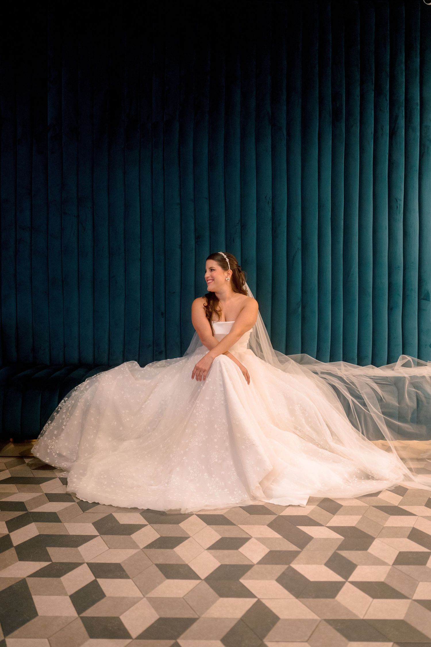 Bride in a voluminous white lace ball gown sits gracefully against a teal velvet backdrop on a geometric tiled floor.