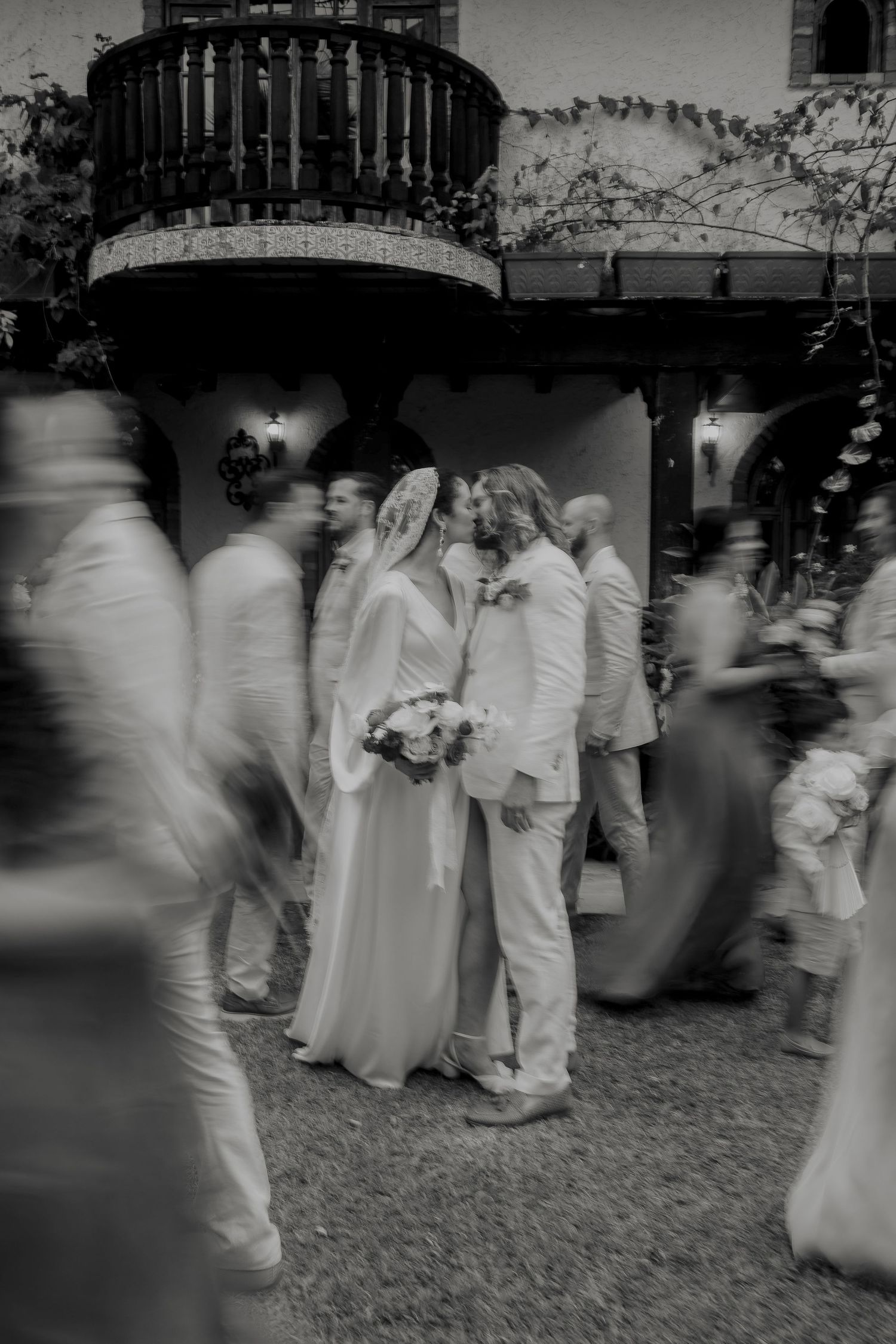 Black and white artistic wedding photo of couple kissing amid blurred moving guests in courtyard.