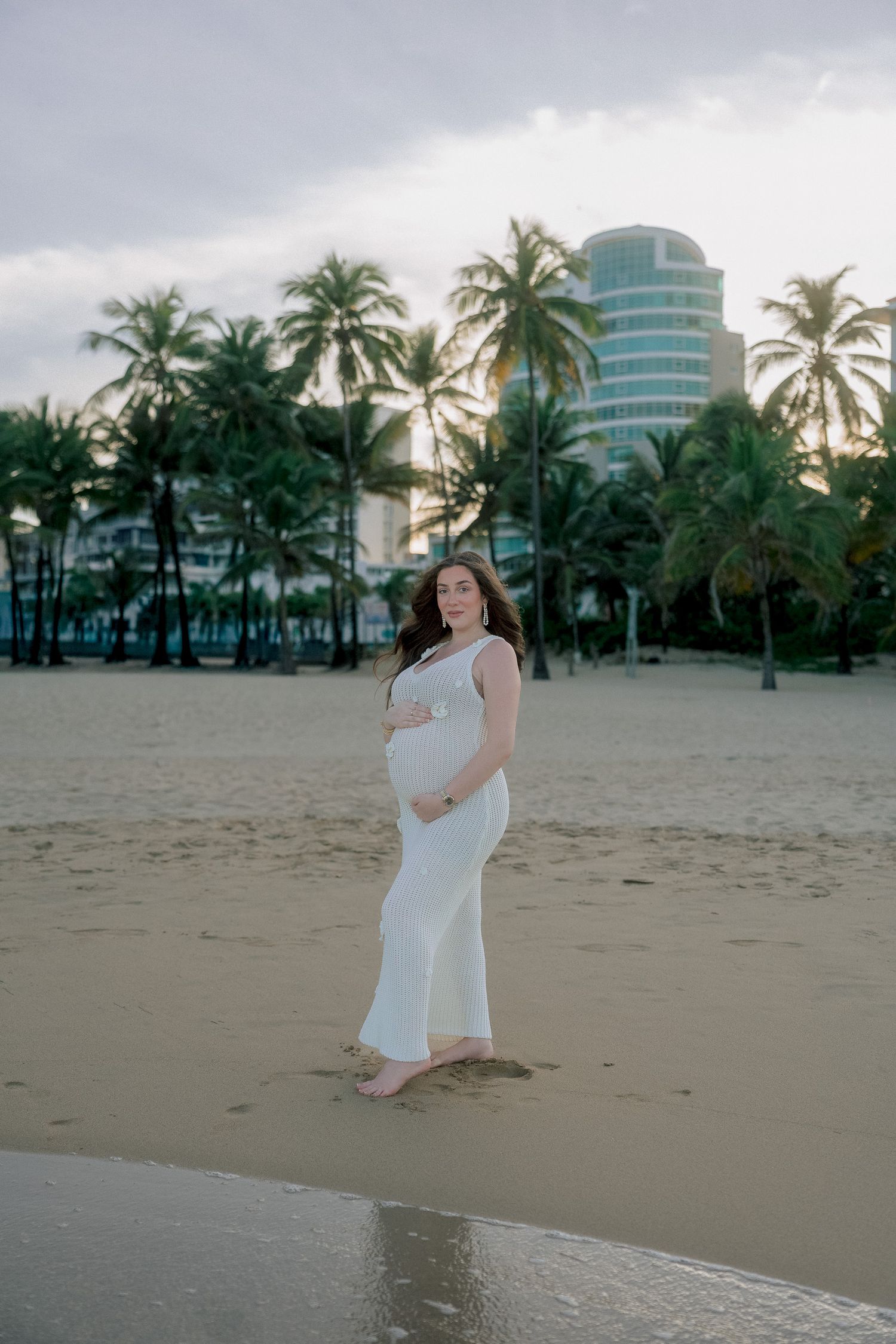 Pregnant woman in white dress on sandy beach with palm trees and building at sunset.