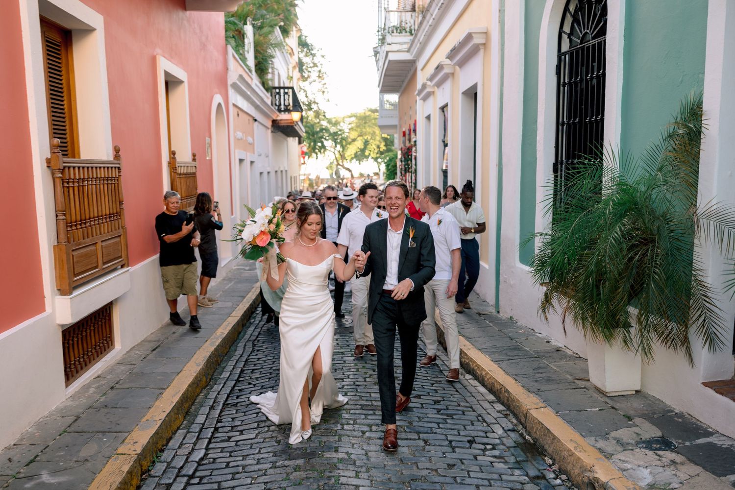 Bride and groom walk cobblestone street in Old San Juan, Puerto Rico, surrounded by colorful buildings and wedding guests.