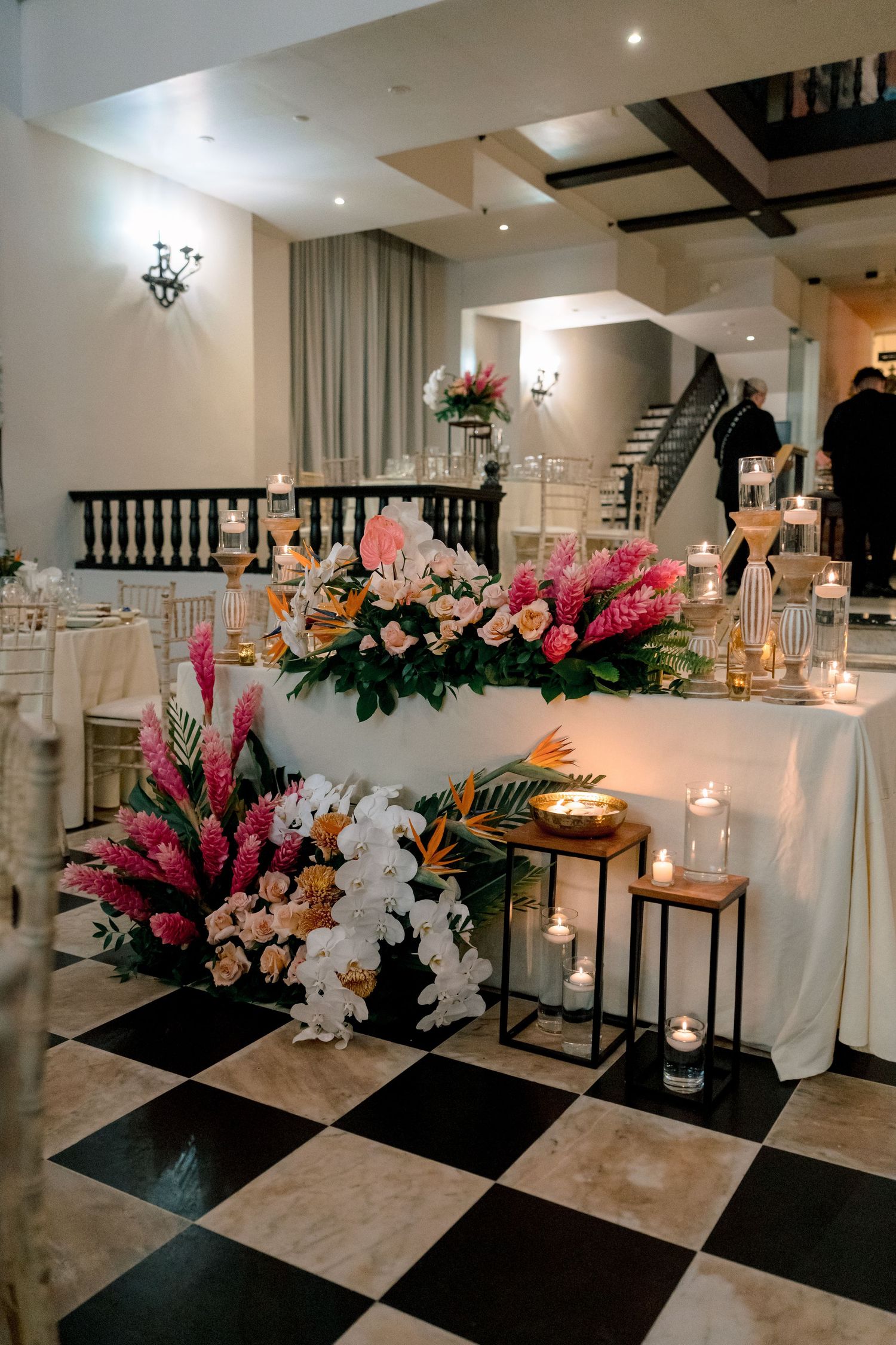 Luxurious wedding reception table adorned with tropical pink flowers, orchids, and candles on checkered floor.