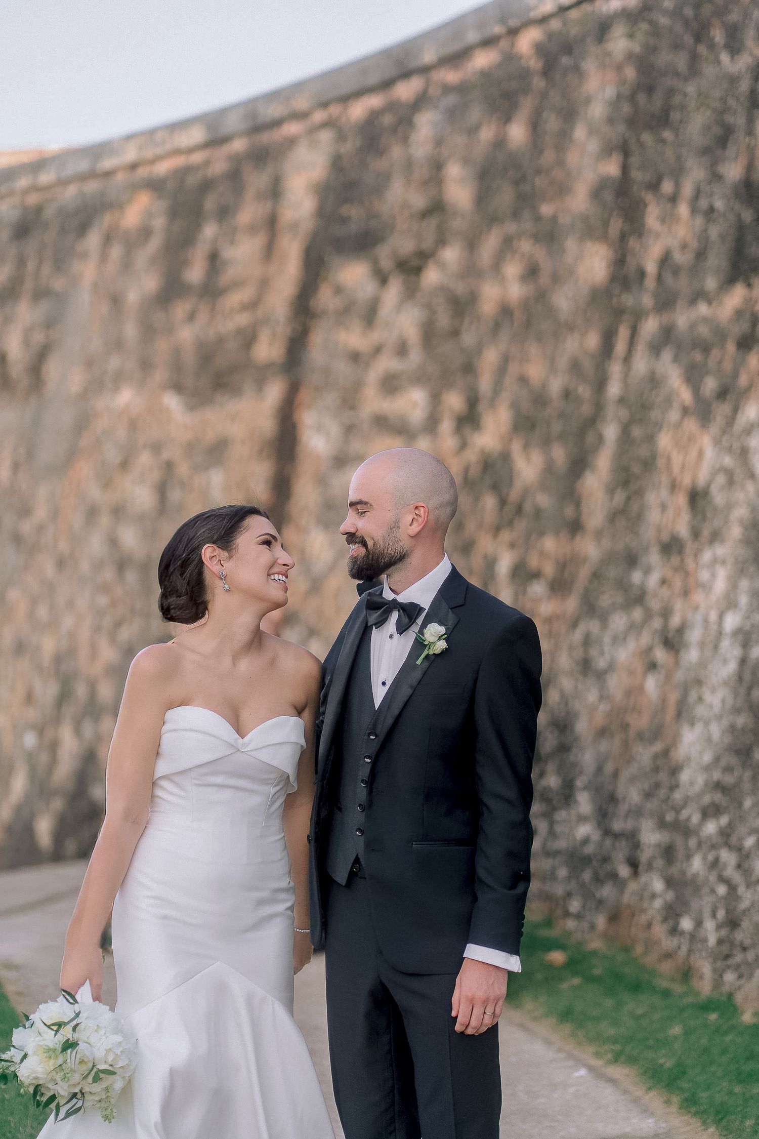 Bride and groom smiling at each other near historic stone wall outdoors.