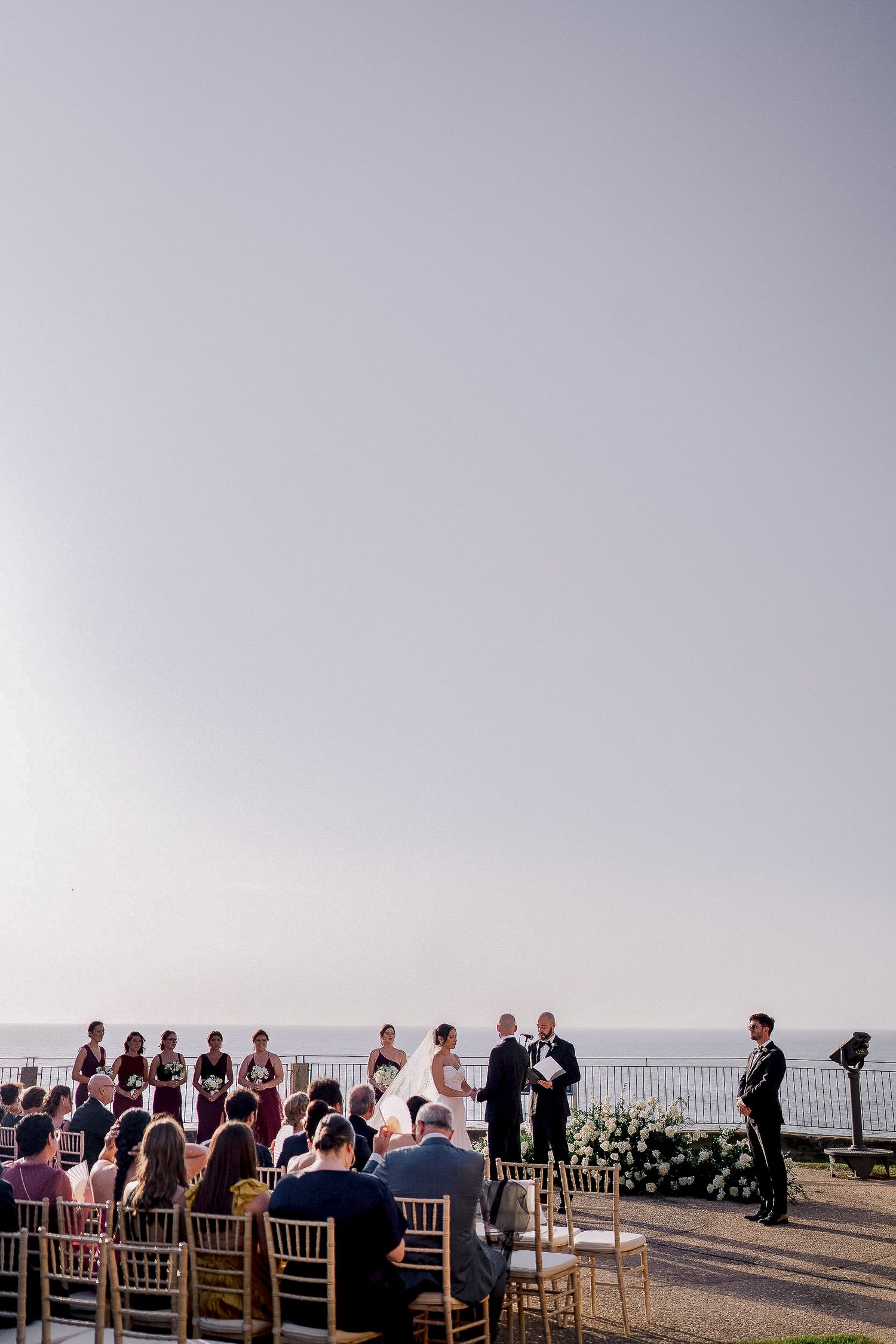 Outdoor wedding ceremony overlooking the ocean with guests seated in golden chairs.