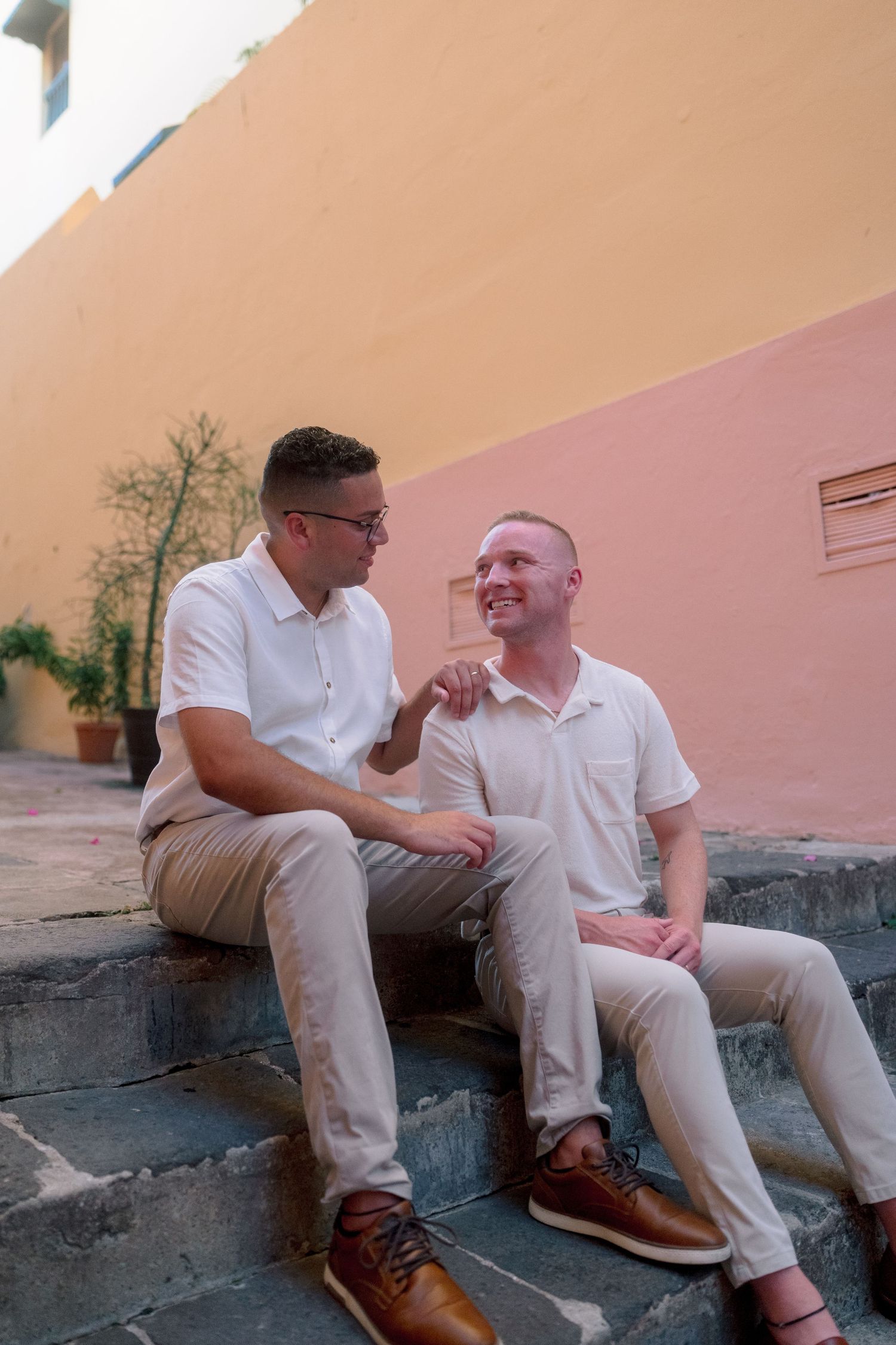 Two men in white shirts and khakis sit on steps, smiling at each other against a warm-toned wall.