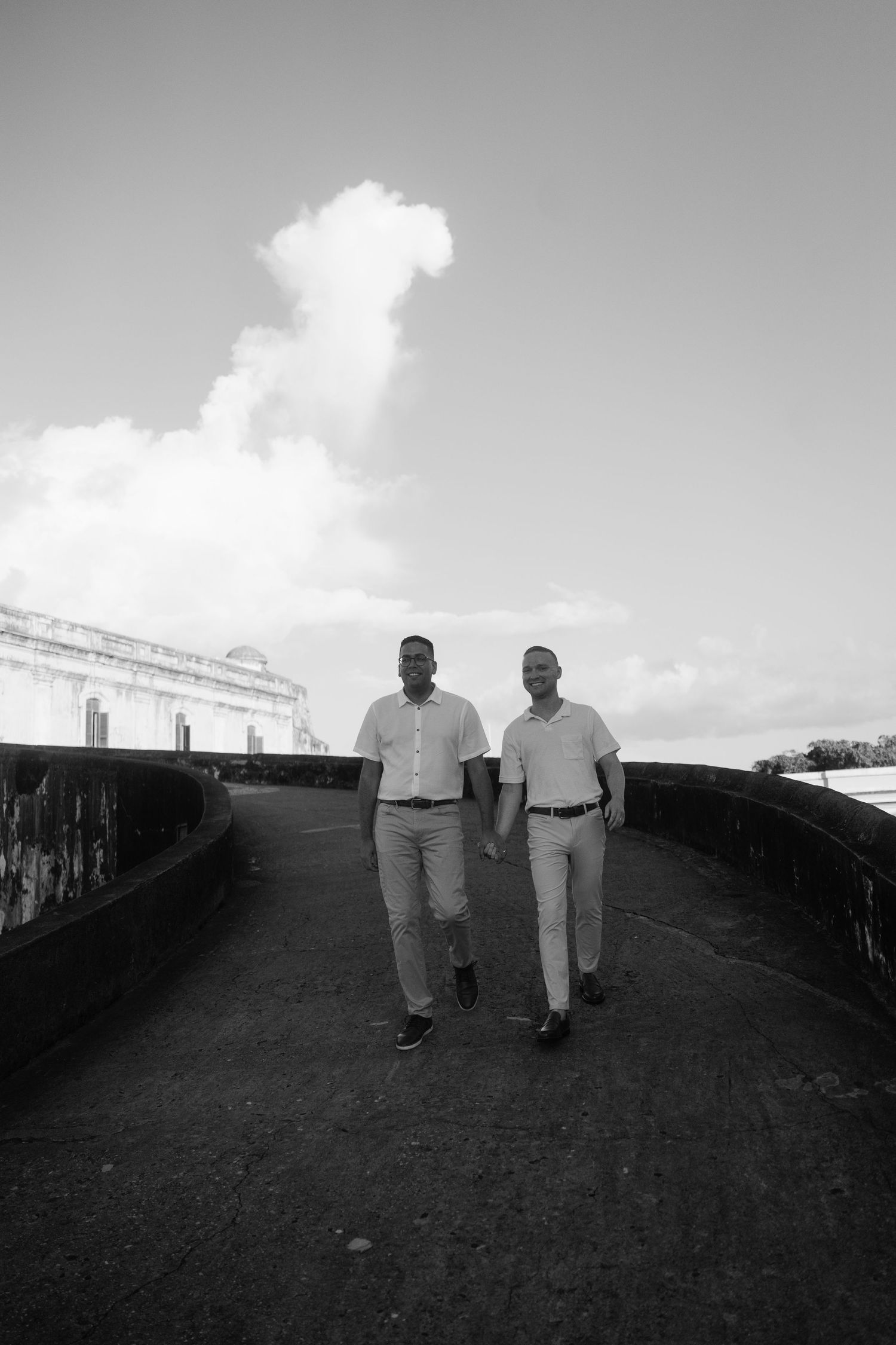 Two men in dress shirts and slacks walk together on a stone path near a historic building under a cloudy sky.