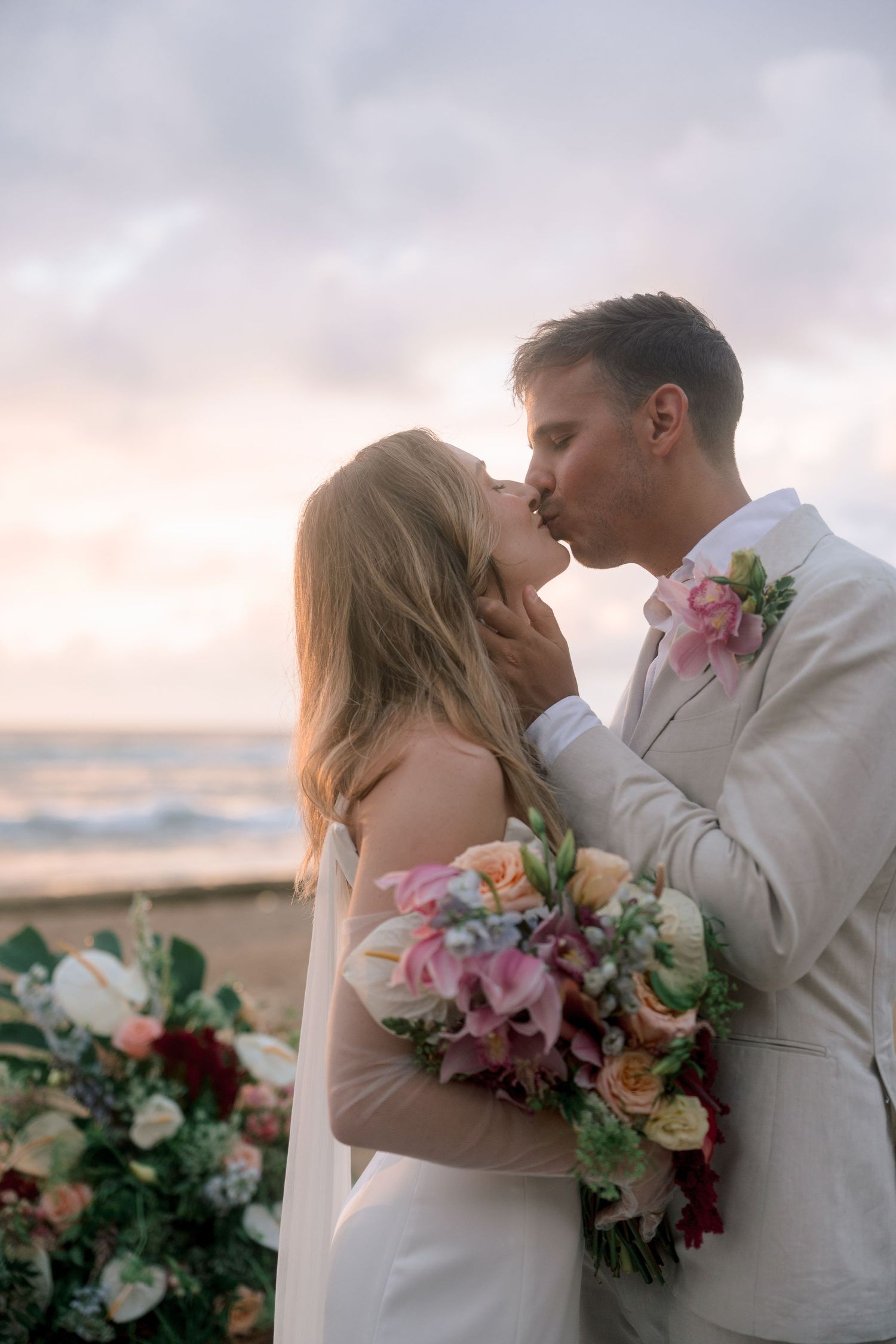 Bride and groom share a romantic kiss on the beach at sunset, with colorful floral arrangements nearby.