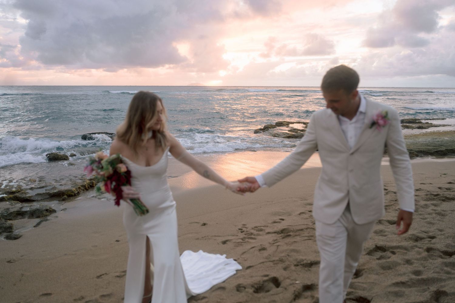 Bride and groom hold hands on a sandy beach at sunset, waves crashing behind them in a romantic wedding photo.