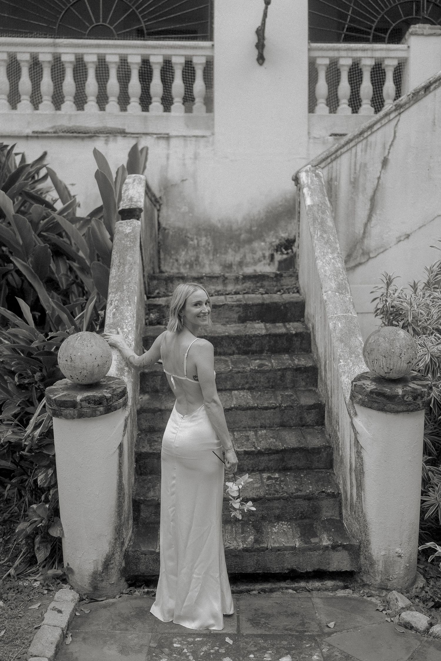 Black and white photo of a bride in a satin gown holding flowers at the base of a vintage stone staircase.