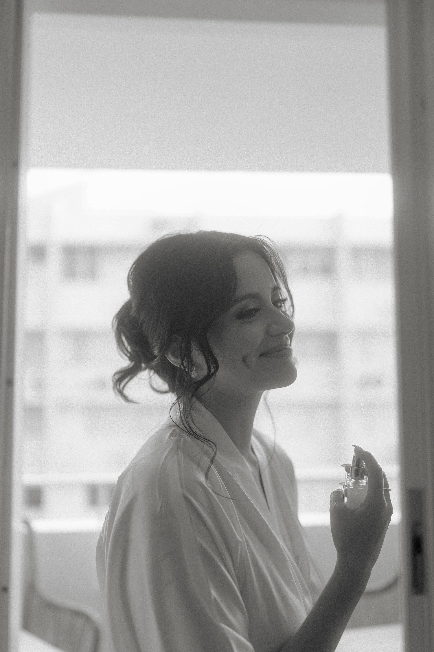 Black and white photo of a smiling bride in a white robe holding perfume by a bright window.