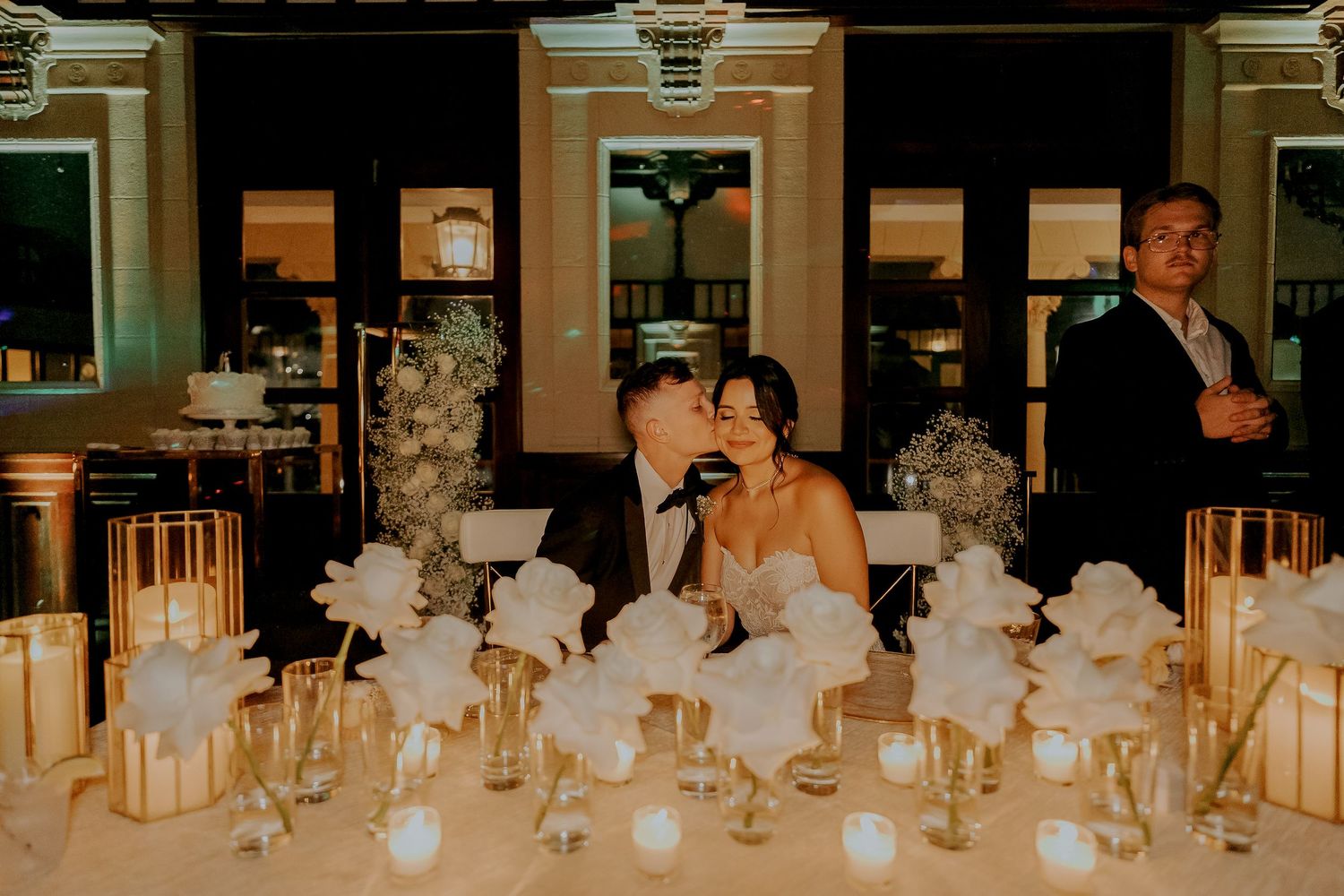 Bride and groom share a kiss at elegantly decorated sweetheart table with white floral centerpieces.