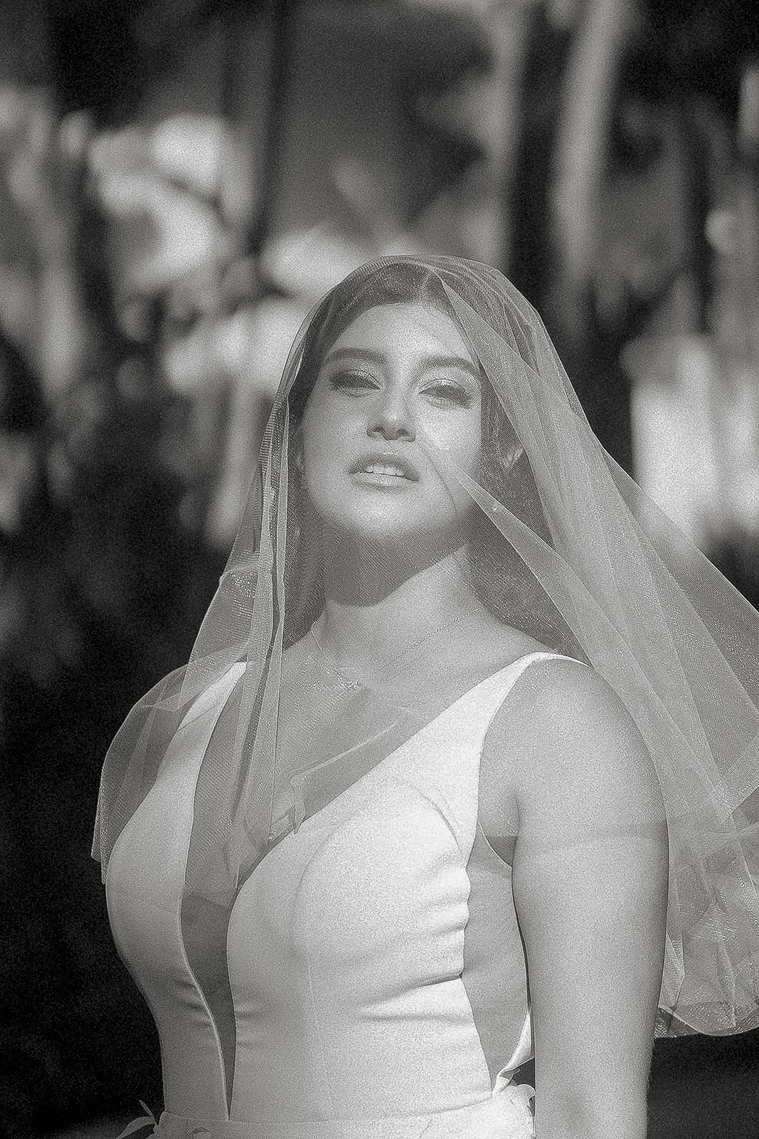 Black and white portrait of a bride wearing a flowing veil, looking upward in an outdoor setting.