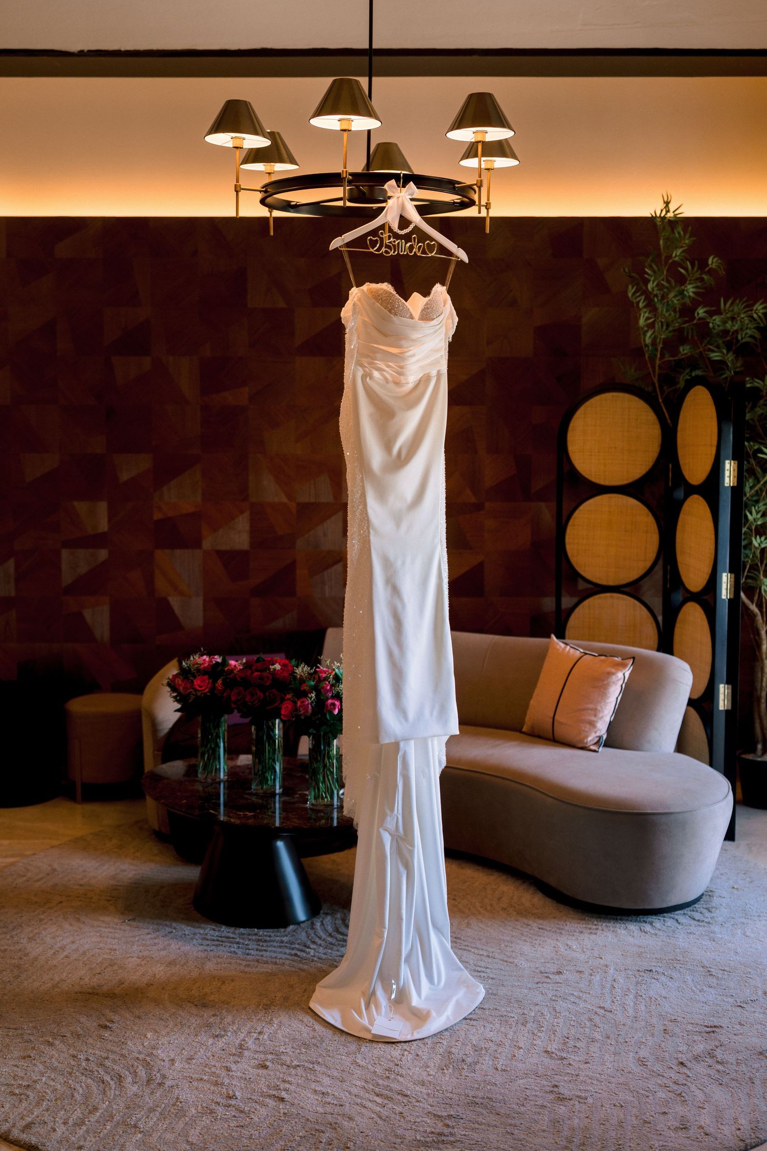 Sleek white satin wedding gown hanging from chandelier in stylish hotel suite.